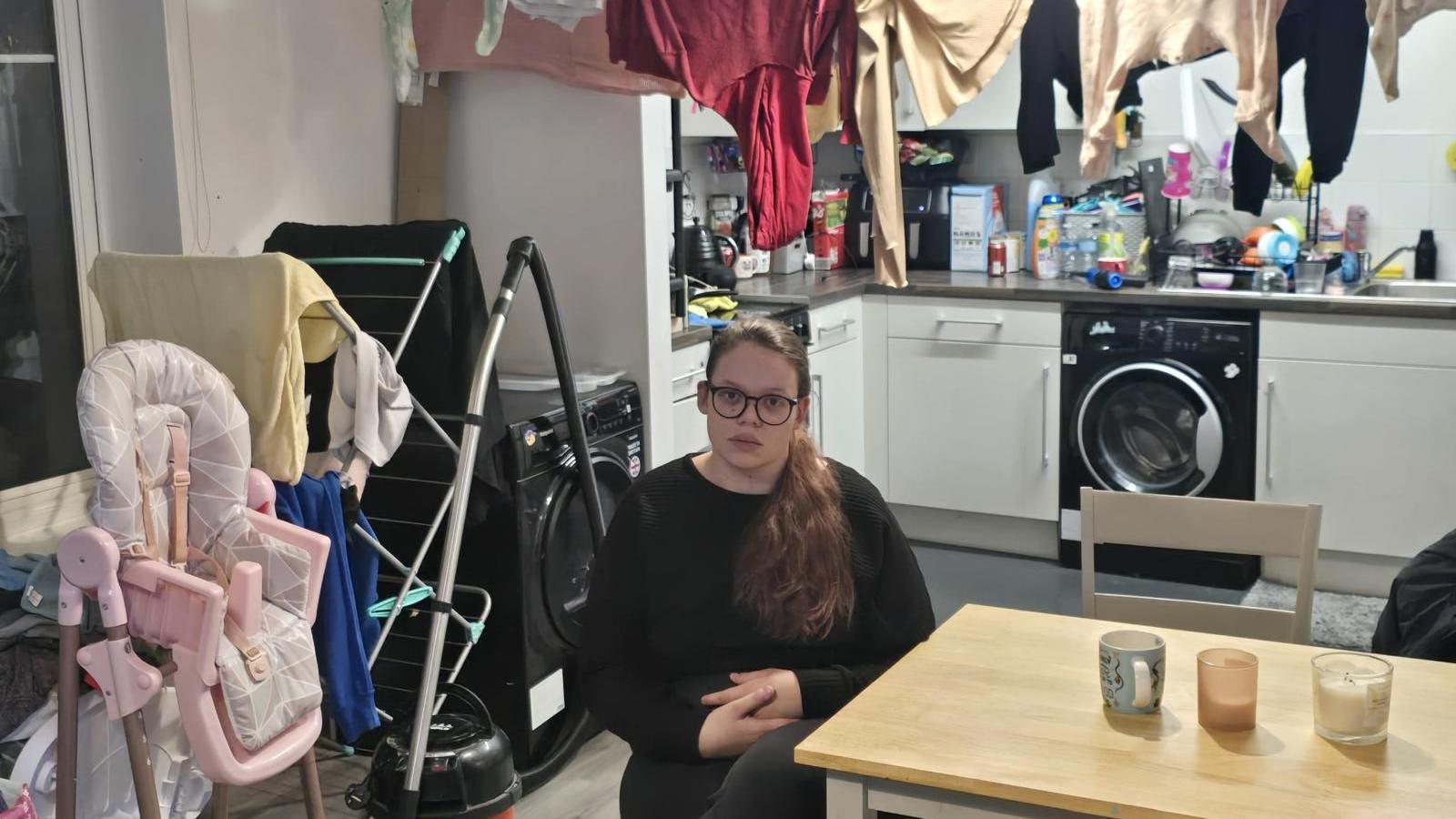 A woman in a kitchen. Laundry is hung above her head. The room looks crowded with items. 