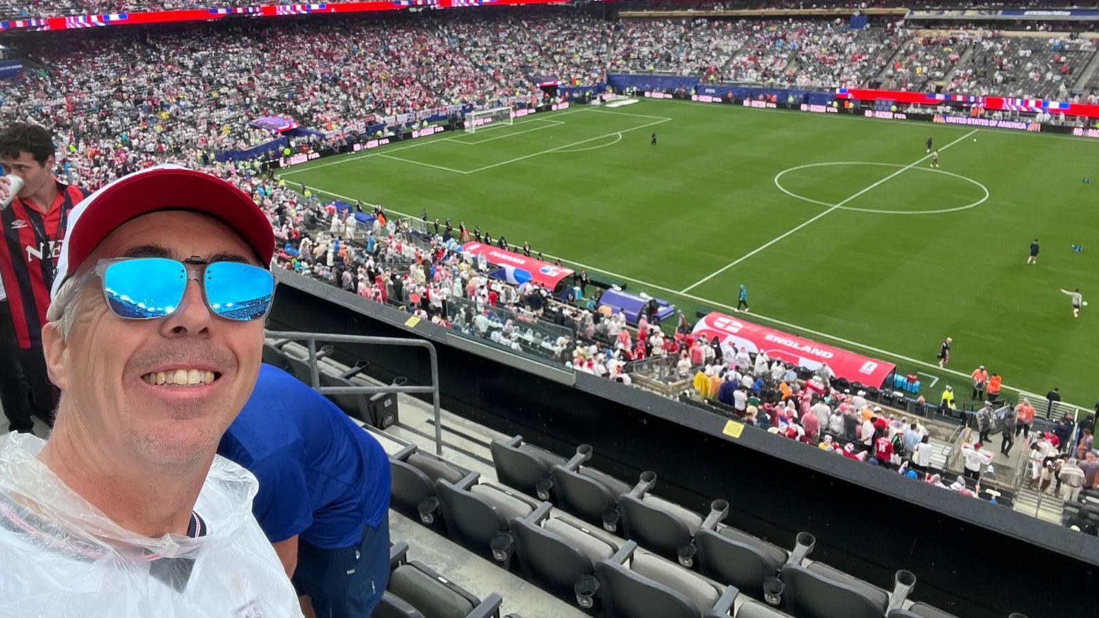 Kevin Craig, wearing a cap and sunglasses, smiles in a selfie inside a packed stadium at the World Cup, with the football pitch in the background. "England" and "Panama" are written over the two team dugouts.