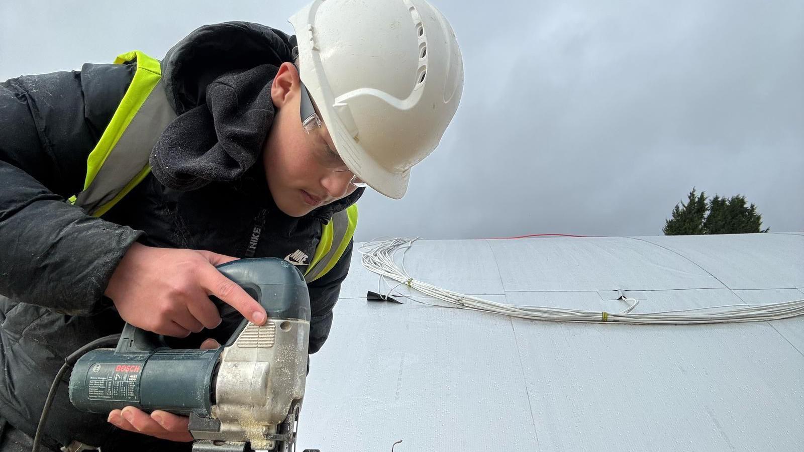 Jack busy at work, wearing a hard hat, hi-vis vest over a black puffer jacket.  He is using an electric jigsaw appliance 