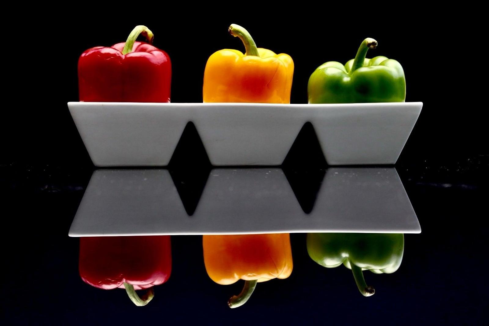 Three peppers in a white bowl reflected on a shiny surface, pictured against a black background