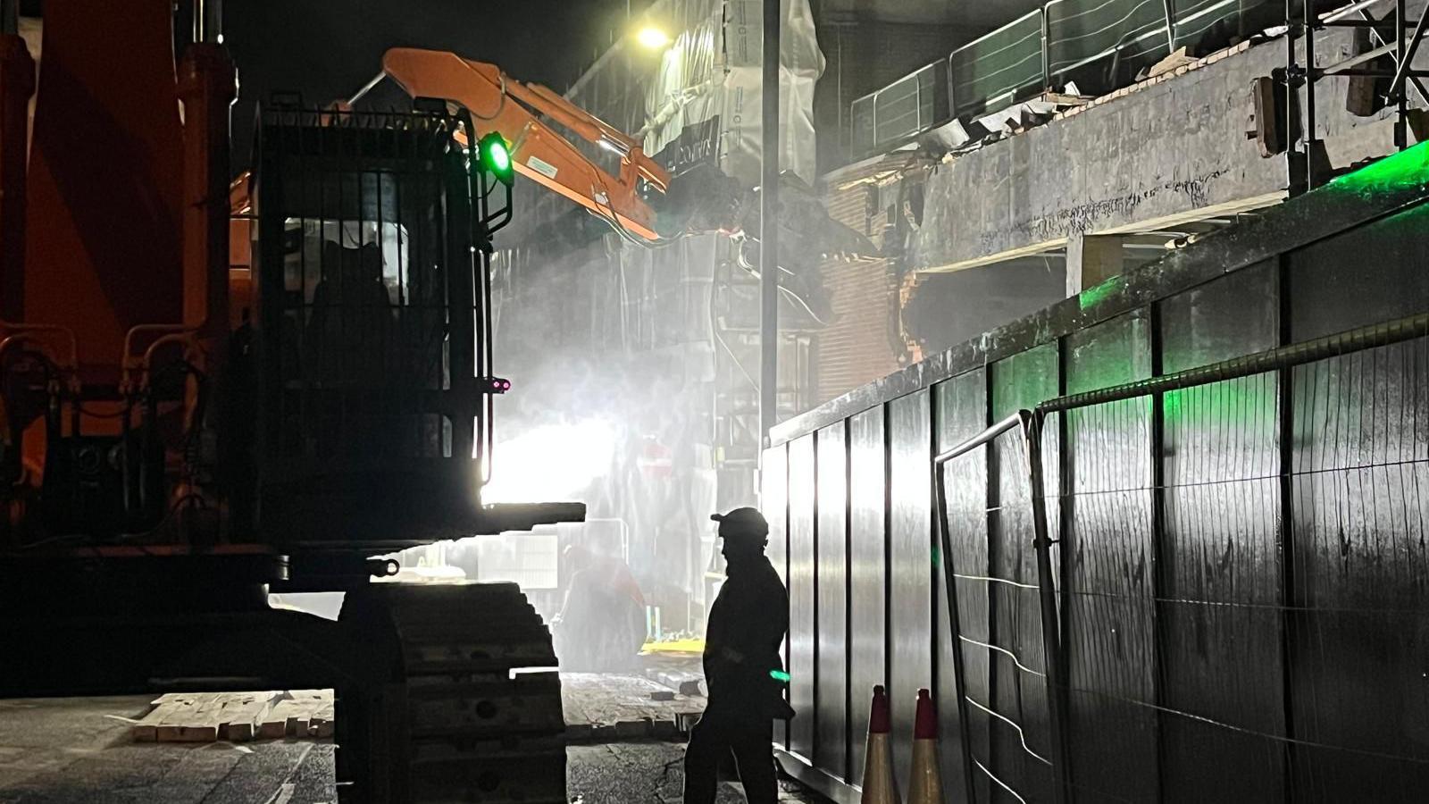 The silhouette of a man in a hard hat standing near heavy machinery being used to bring down a wall as part of the demolition.