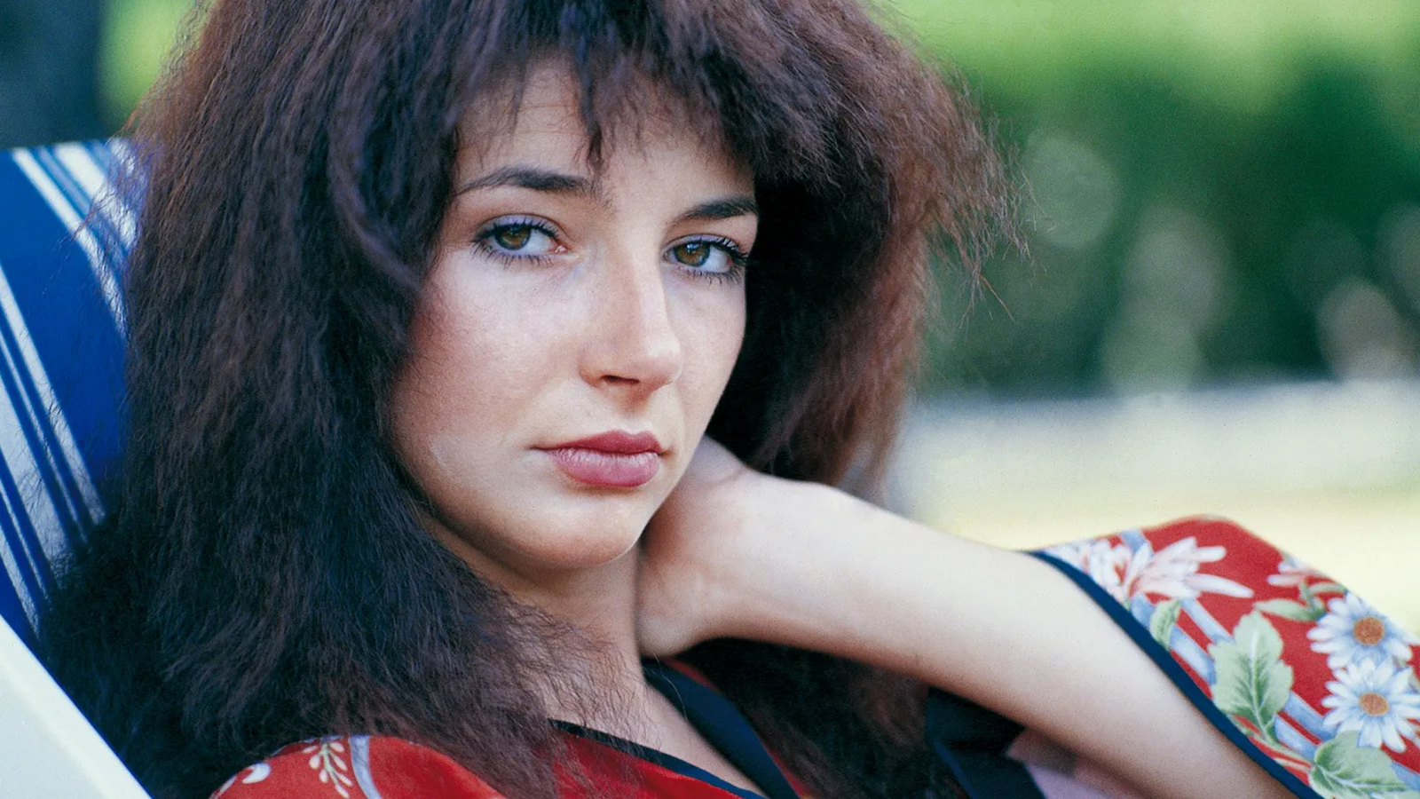 Kate Bush, with a hand resting on her neck, looks directly into the camera while sitting on a chair outdoors.