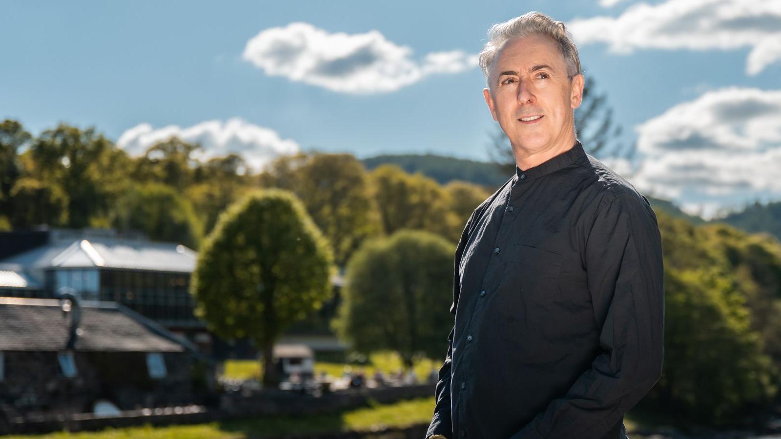 Actor Alan Cumming, a grey-haired stylish man in a black shirt smiles while looking off to the left as he stands with Pitlochry Festival Theatre in the background - it is a building with a lot of windows set in beautiful green countryside.