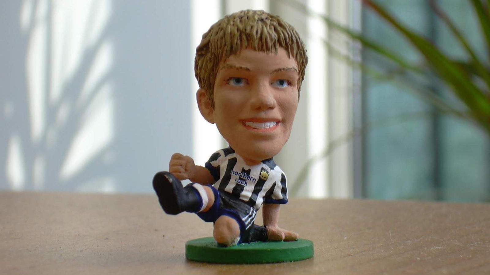 A close up of his model. It has short blond hair and blue eyes and is wearing a black and white Newcastle strip. The pose is of James sliding on one knee, one foot and arm raised as if making a tackle.