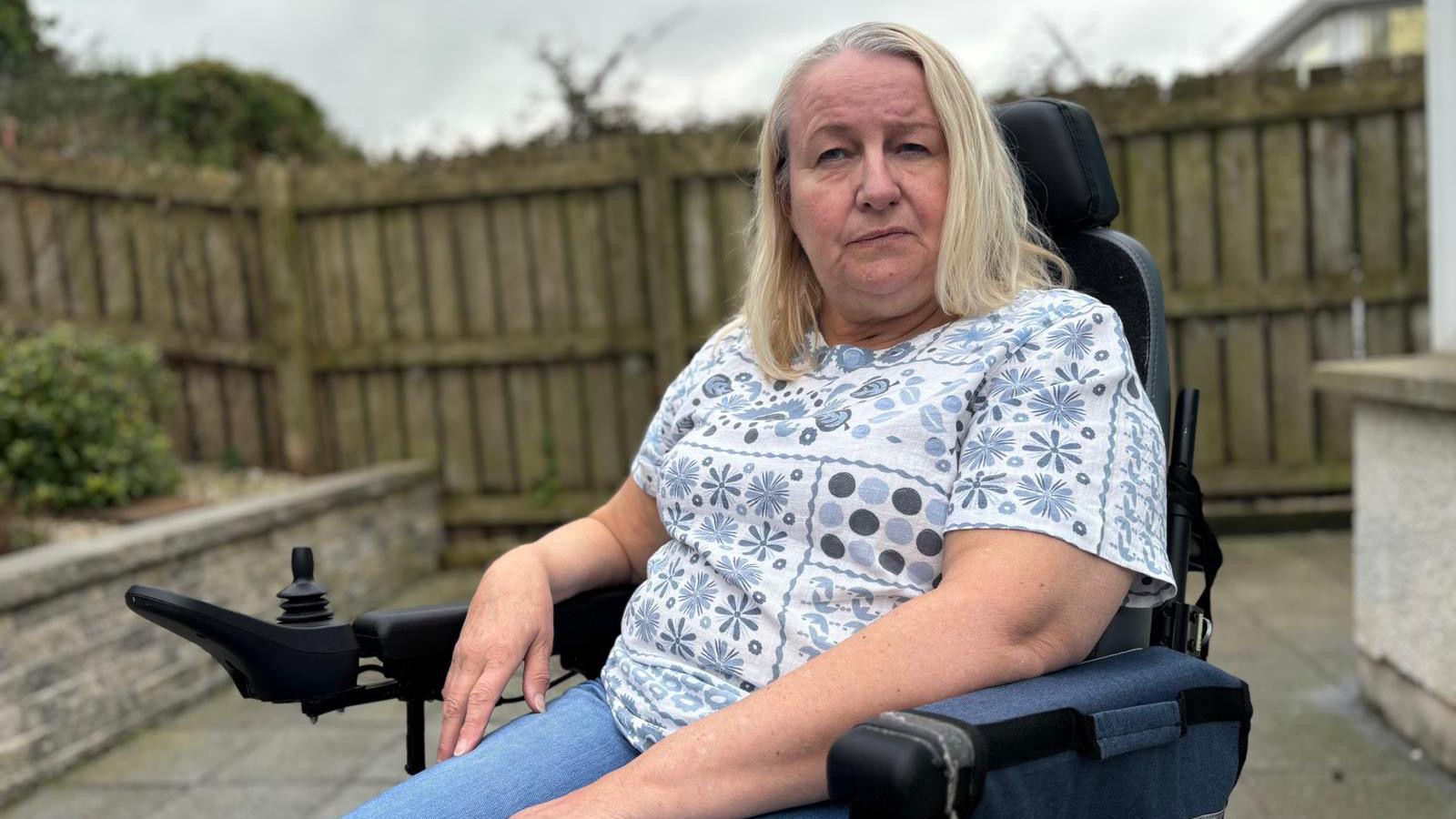 A woman in a motorised wheelchair looks off to the side of the camera. She has light blonde hair and is wearing a blue and white patterned top and light blue jeans. The background is out-of-focus, but you can see a wooden fence and paving stones behind the woman.