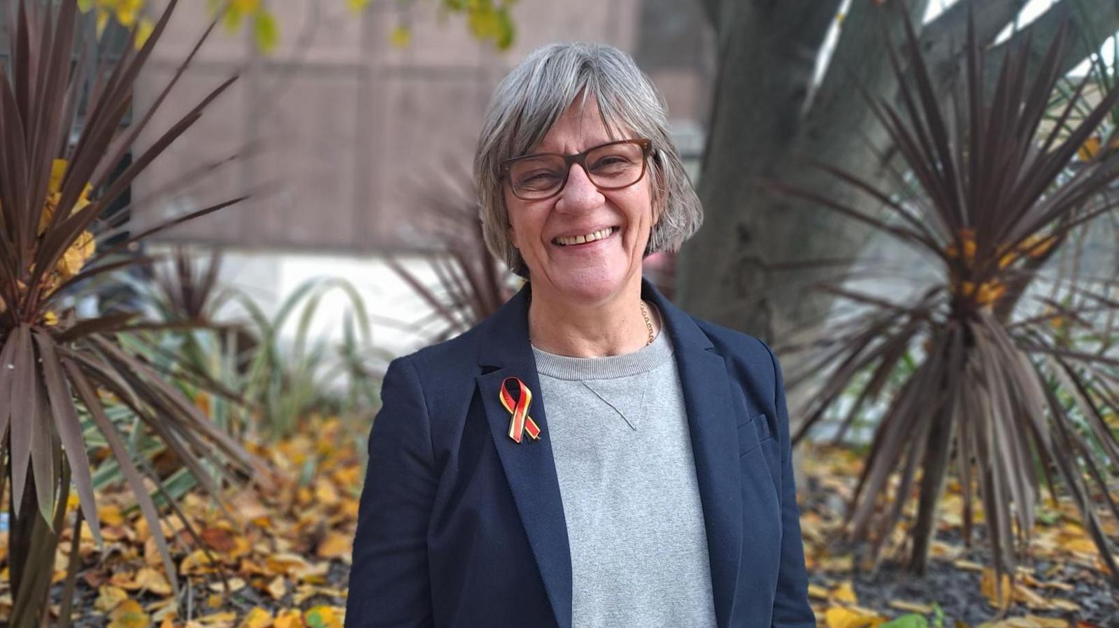 Samantha May, helpline manager, looking at the camera and smiling in a garden with plants and trees in it. She is wearing a blue blazer and a grey top underneath.