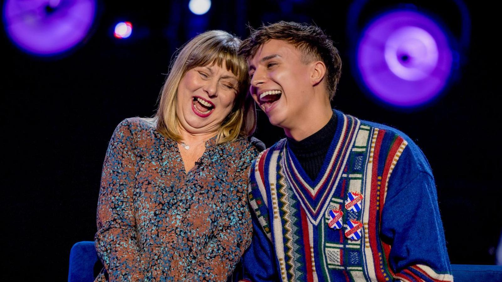 Paul Greaves wears a blue jumper with a red, blue and white pattern. They are wearing three badges. They are sat beside their mother Diane who is wearing a patterned blouse. She has blond hair and the pair are laughing.