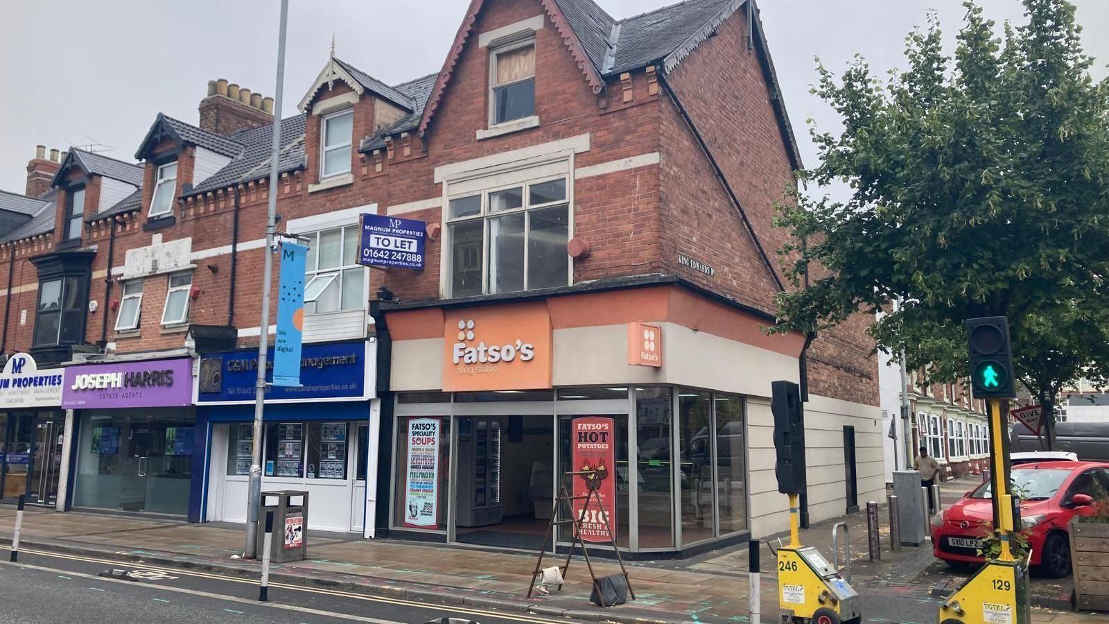 Fatsos located on Linthorpe Road. A building sits on the corner of a road, with large glass frontage and an orange sign over the entrance which reads ' Fatso's' in white lettering. It is in a row of shops and a road is in front of it with temporary traffic lights.