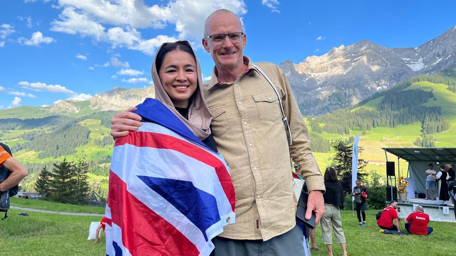 Fatima Painda draped in a Union flag and standing next to her coach, Paul Harkins. There are mountains in the background