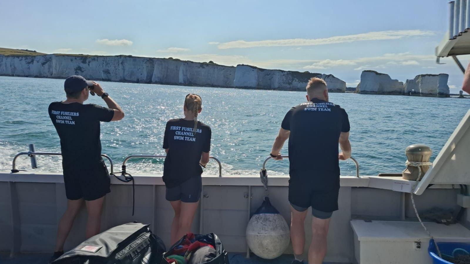 Three adults stand by the side of a boat watching the White Cliffs of Dover as they pass. On the back of their shirts, it reads "first fusiliers channel swim team."