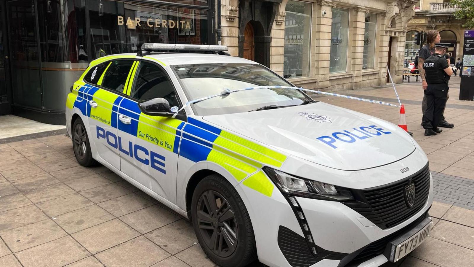 A police car parked on the pavement. Police tape has been attached to it to help create a cordon in front of a stone building. Two police officers are standing in front of the tape. 