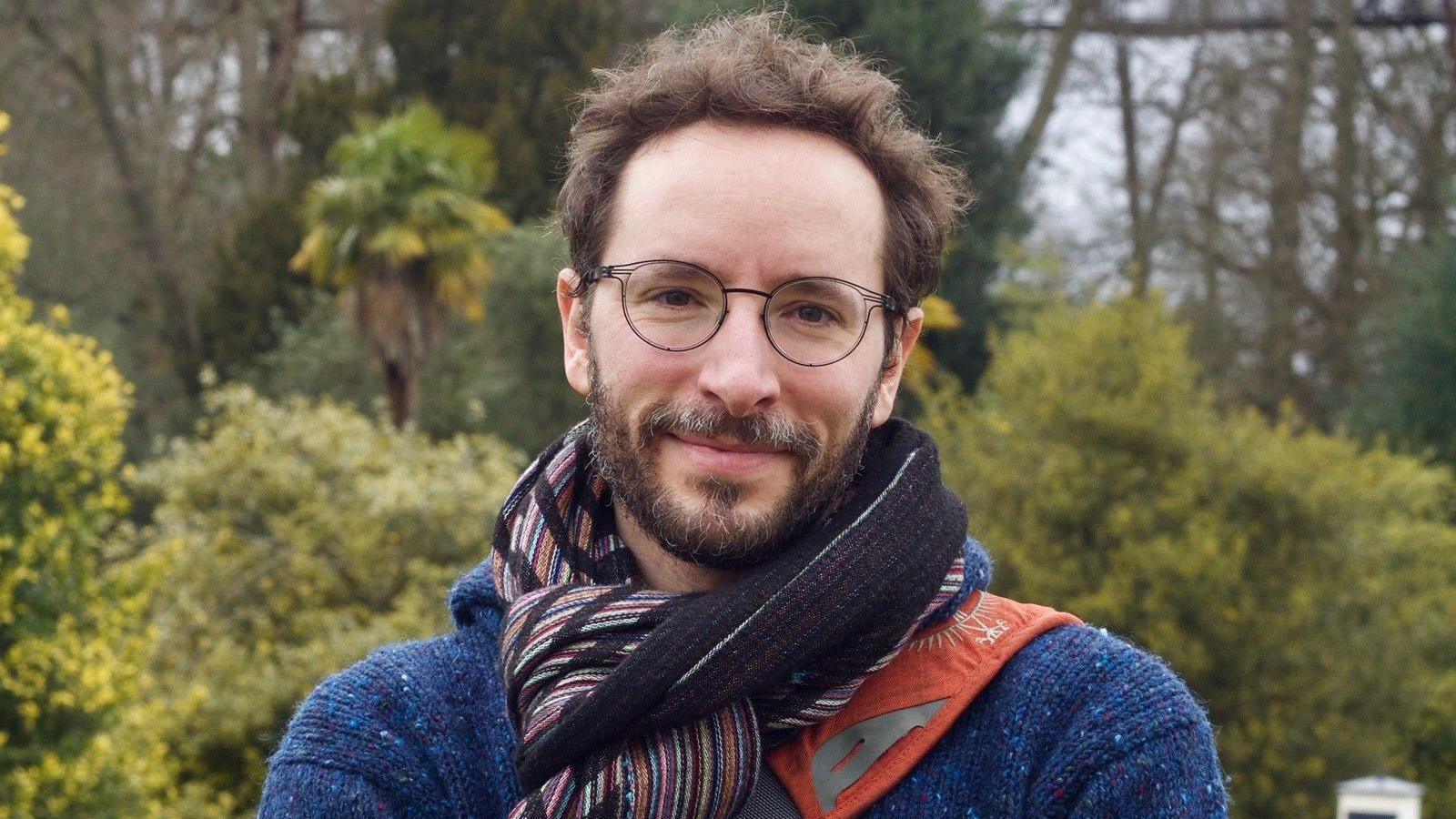 A file image of Oliver Lord, a man with glasses and a beard, wearing a red striped scarf, blue jumper and orange backpack. He is standing in front of some trees.