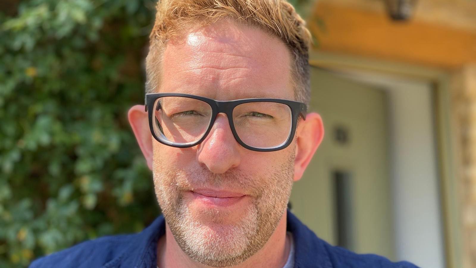 A head and shoulders photograph of Jonathan Baynes who wears a blue shirt and black-rimmed glasses and has light-brown hair, cropped short at the sides