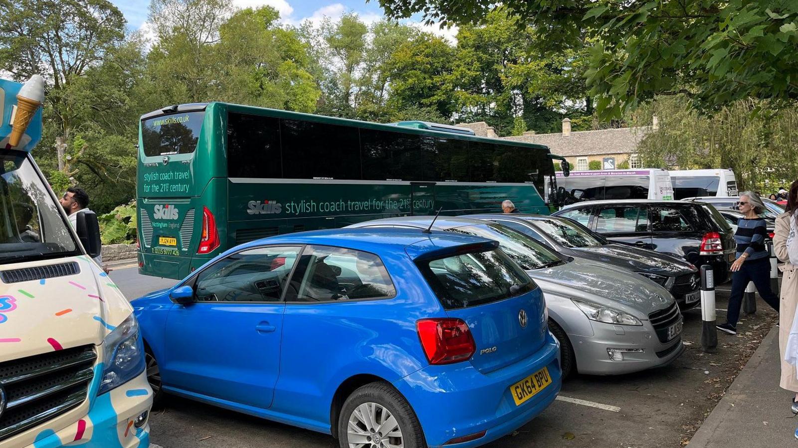 A green coach drives past a row of parked cars and an ice cream van in the Cotswolds village of Bibury. A honey coloured Cotswold stone pub can be seen in the background and there are trees everywhere.