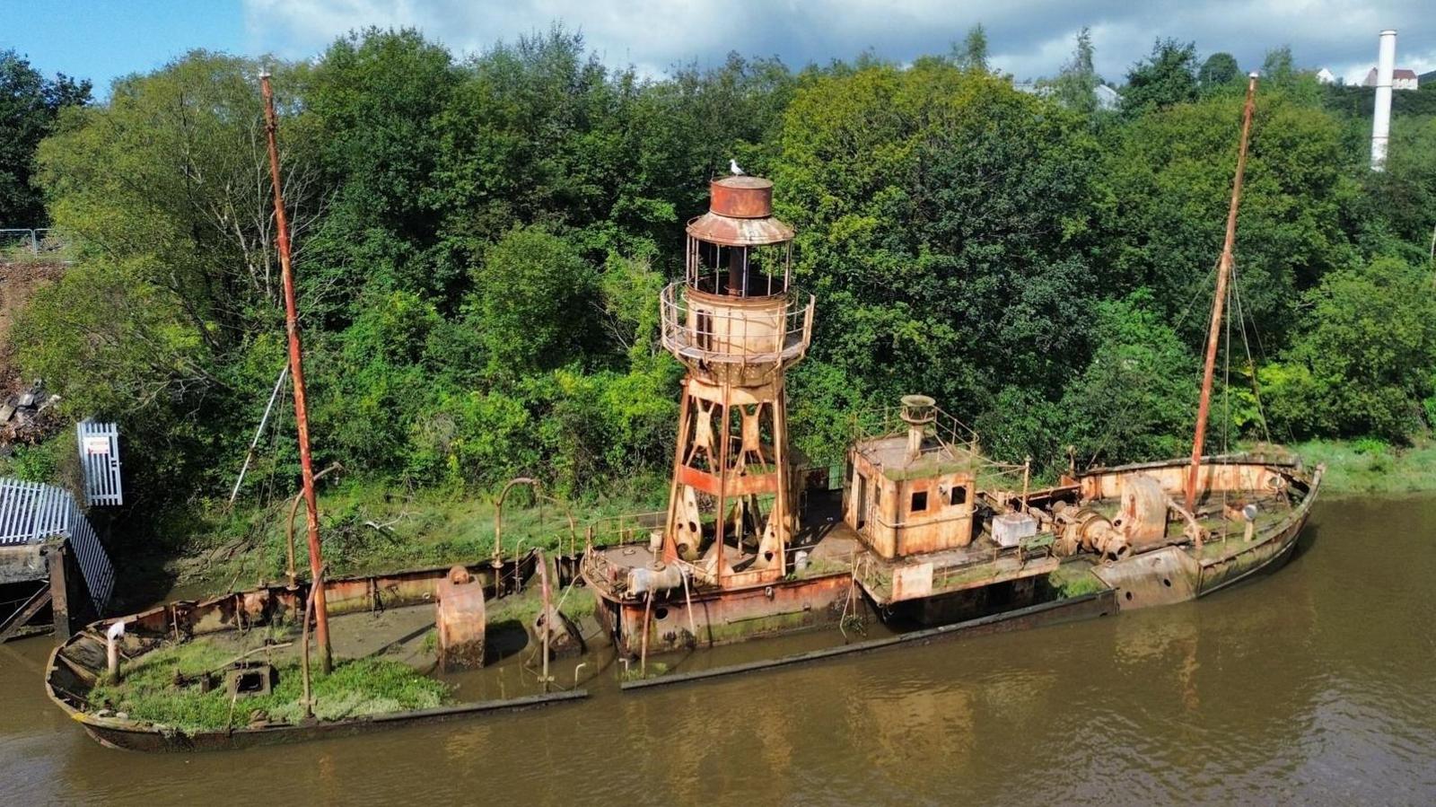 A rusty ship with a light house attached to it. It is slightly leaning, with greenery behind it and muddy water in front.