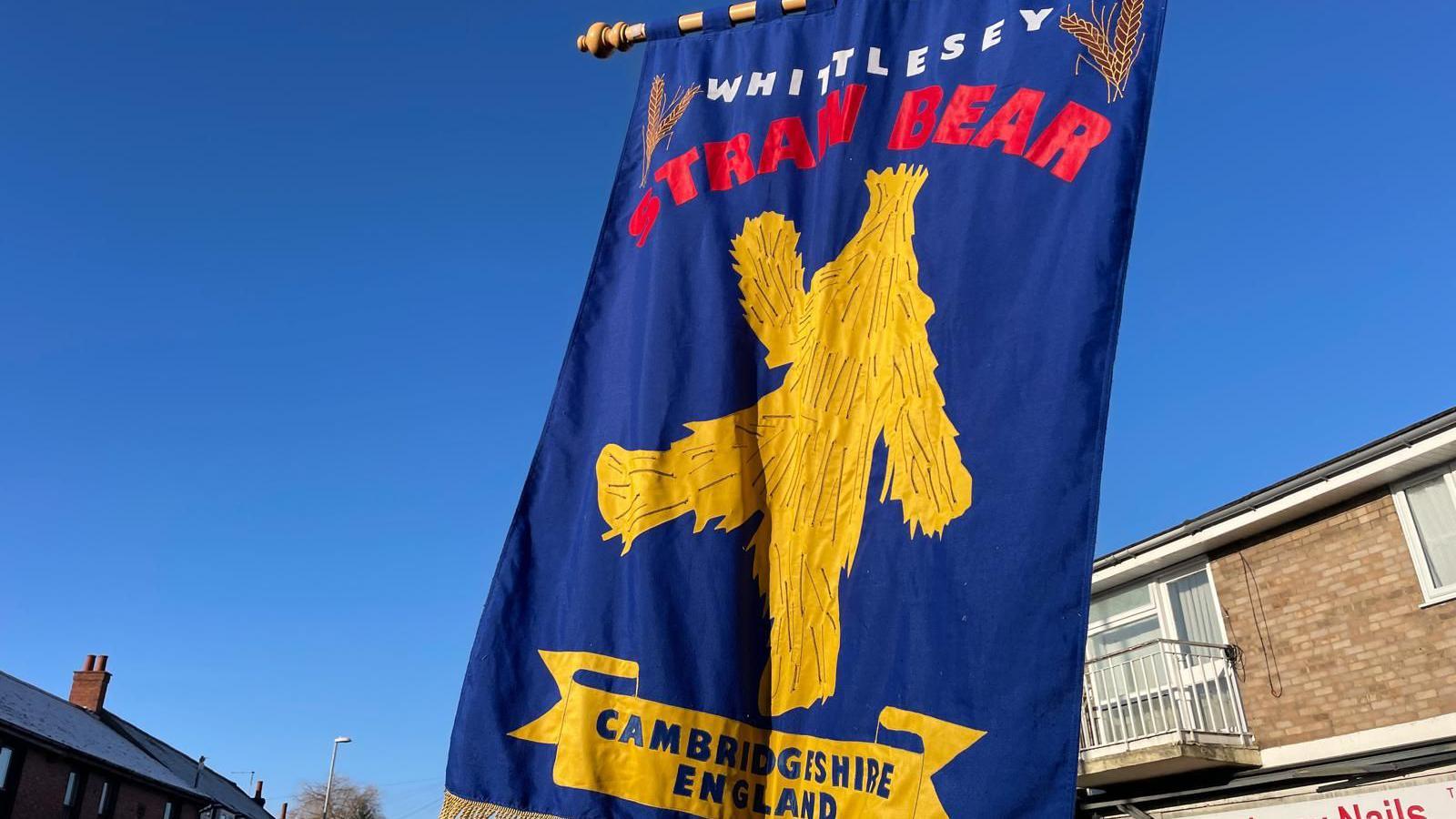 A blue flag which says WHITTLESEY in white writing, a figure of a straw bear and red writing which says STRAW BEAR.