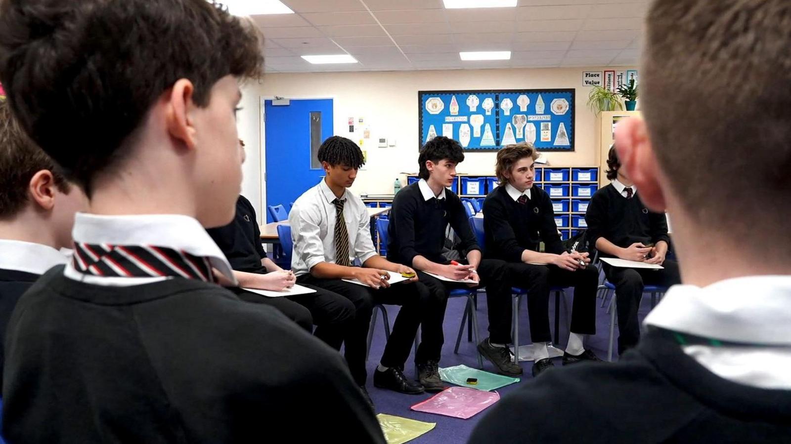 Boys in school uniform sit in a classroom. They are listening to, and looking at, an unseen speaker.