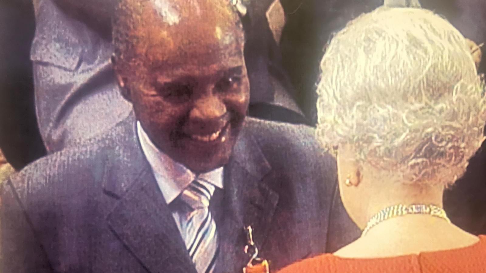 Dr Stephenson smiles as he receives his OBE from Queen Elizabeth II. He wears a dark grey suit and a striped tie, and the medal is partially visible on his lapel. The late Queen can be seen only from behind and is wearing a red or orange dress, with a diamond necklace. 