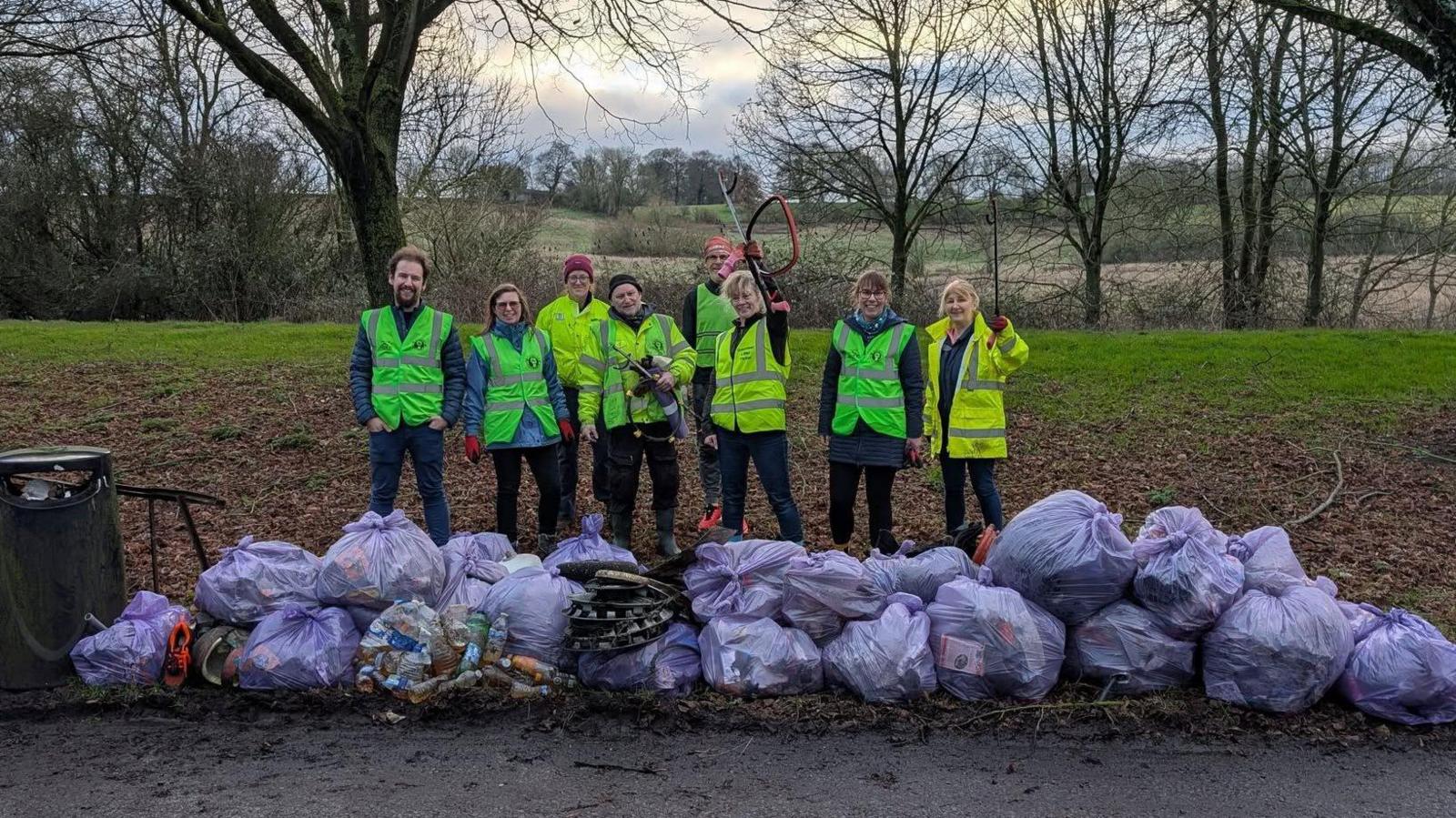 A group of volunteers wearing yellow hi-vis vests standing behind of row of heaped purple rubbish bags next to a path in a green space with a grassy area, trees and fields behind them