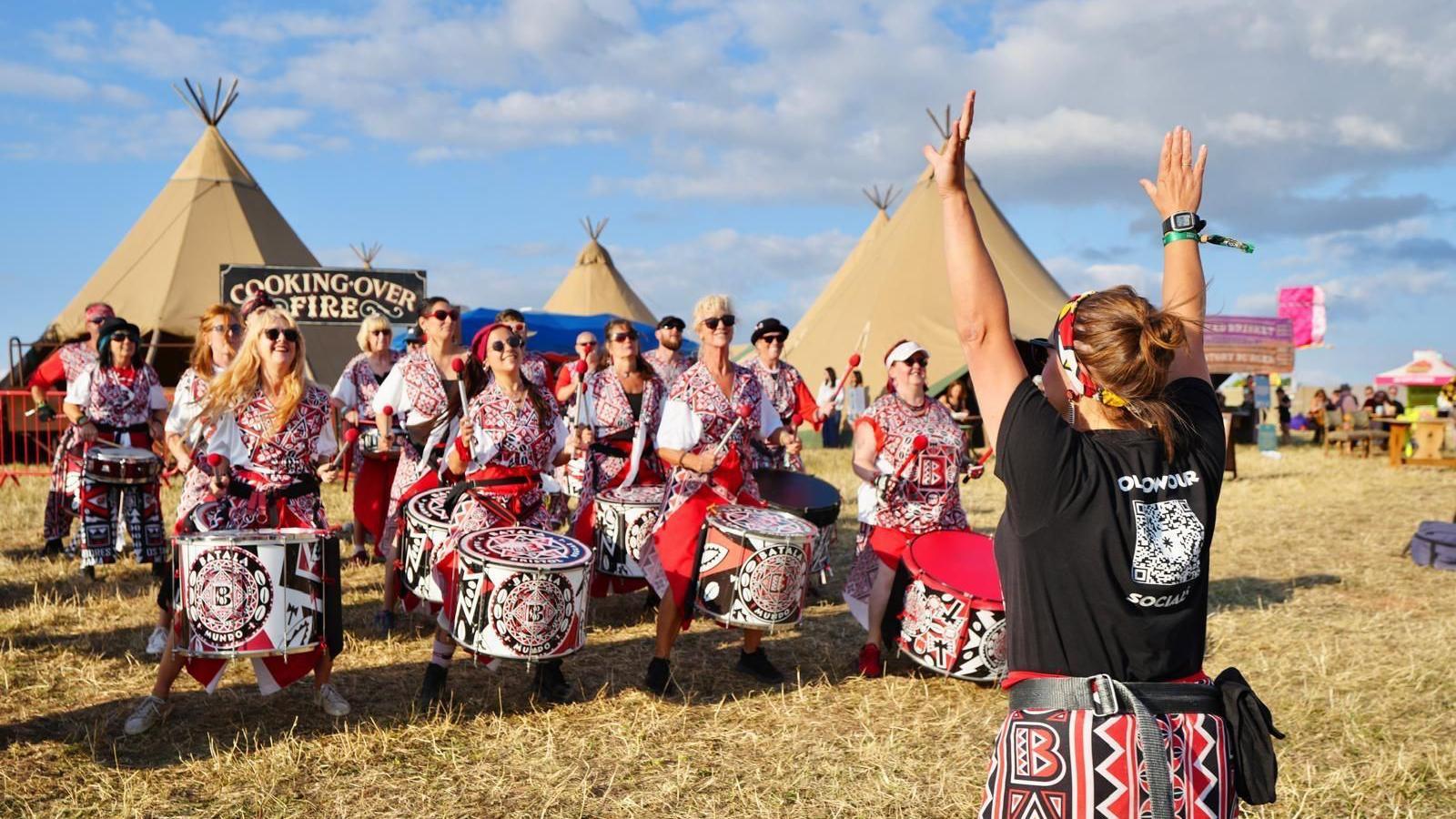 A group of drummers line up on a grassy area near some tepees at the Lake Paradiso festival at Chew Valley Lake. The drummers all have large drums in front of them and are wearing vivid red and black outfits