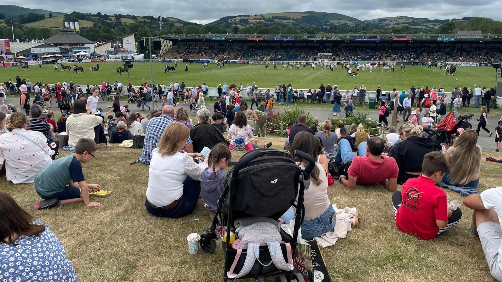 A wide image of the large crowds watching the main ring at the show. There are lots of horses and people in the ring in the background. 