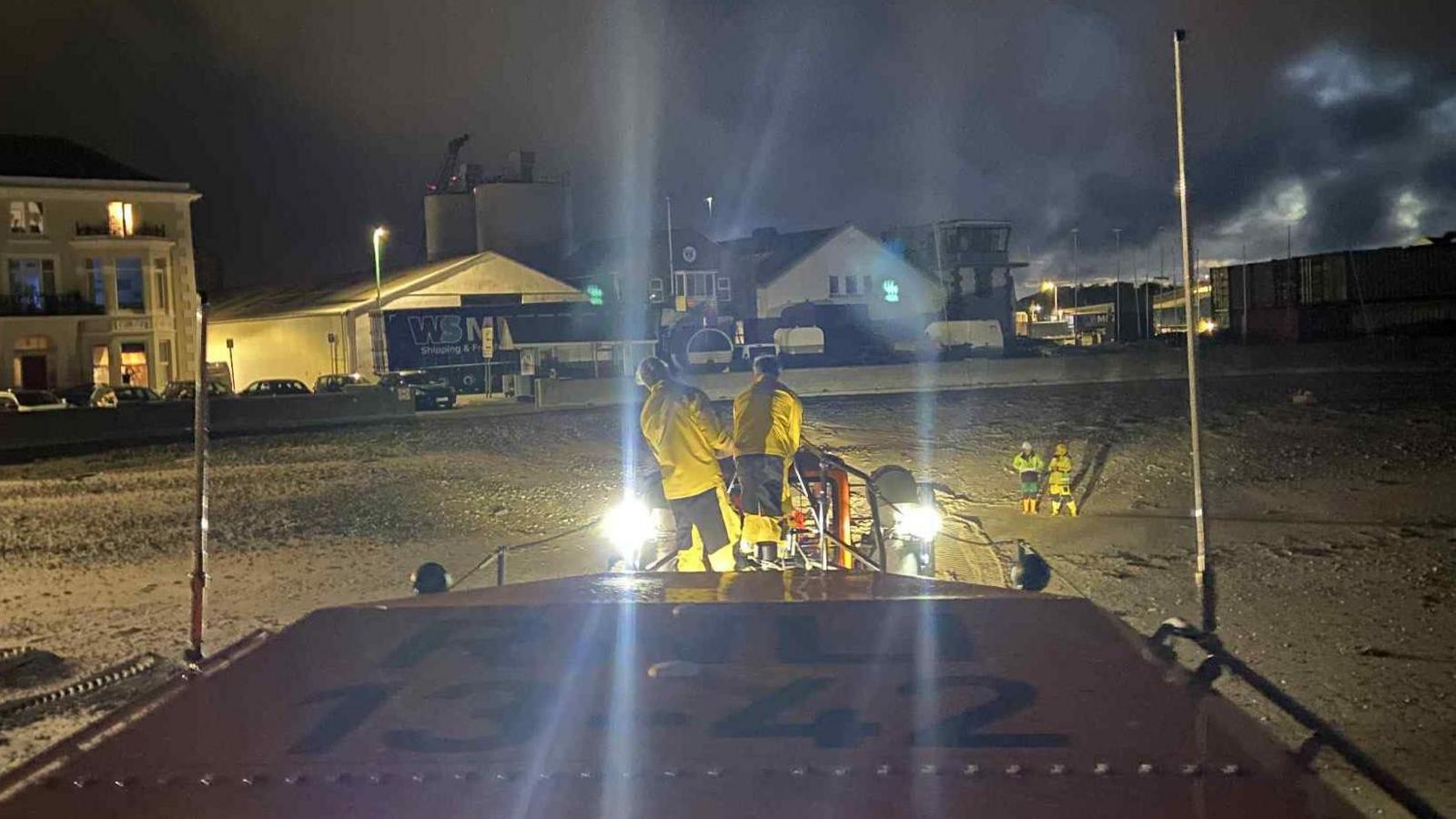 A view from the front of a lifeboat as it returns to shore after dark. Two people in yellow uniforms stand at the front of the vessel, and two others can be seen waiting on the sand.