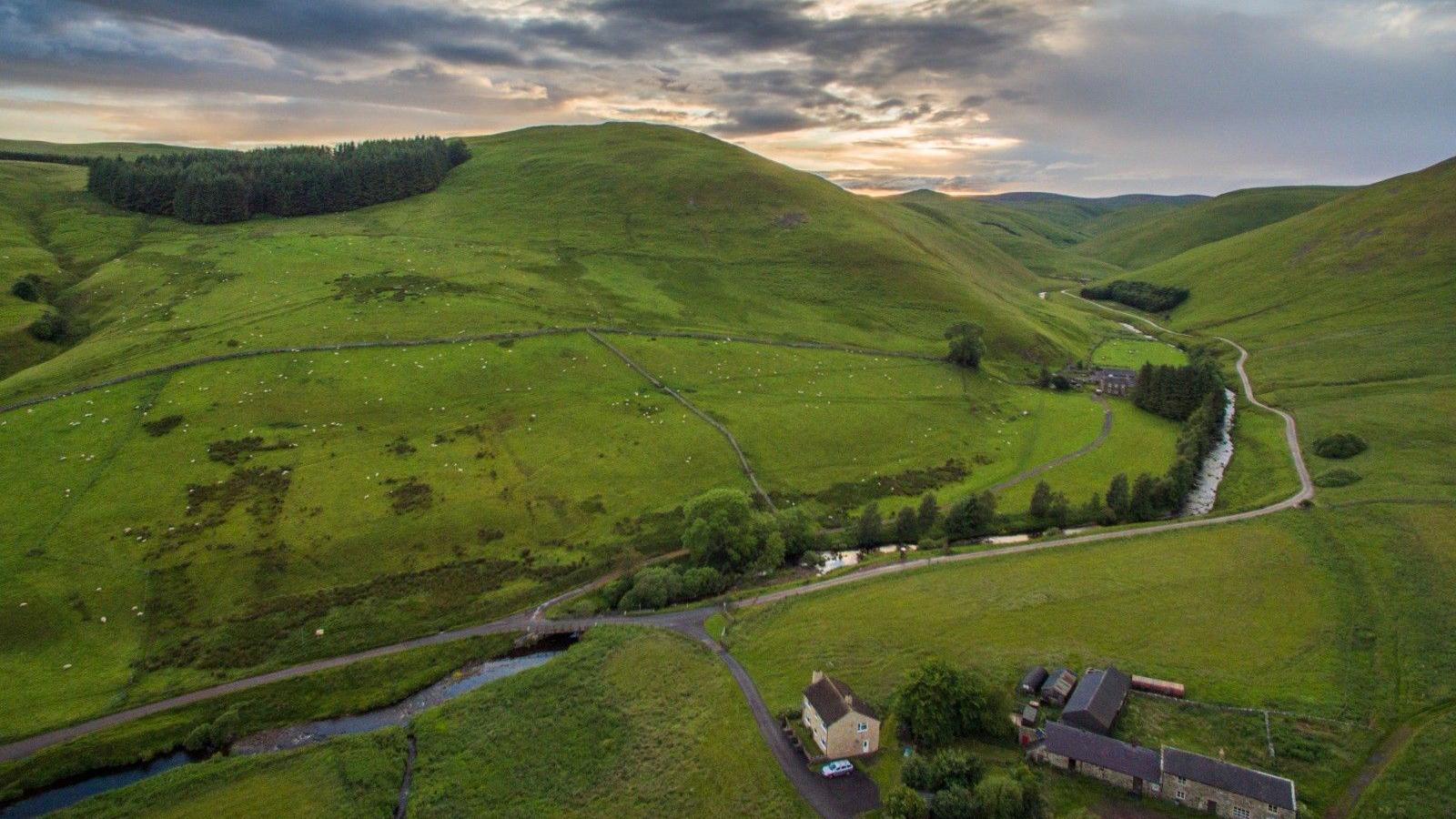 Image shows a green valley taken from the air with a farm in the foreground and a river with a road bending round 