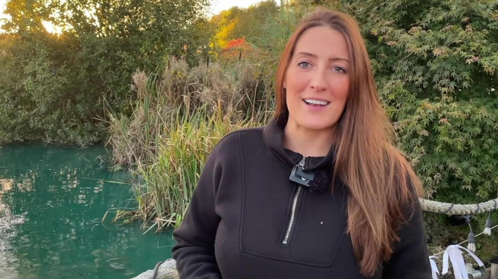 Barbara Ikin has long brown hair, is wearing a black top, and is standing next to a lake