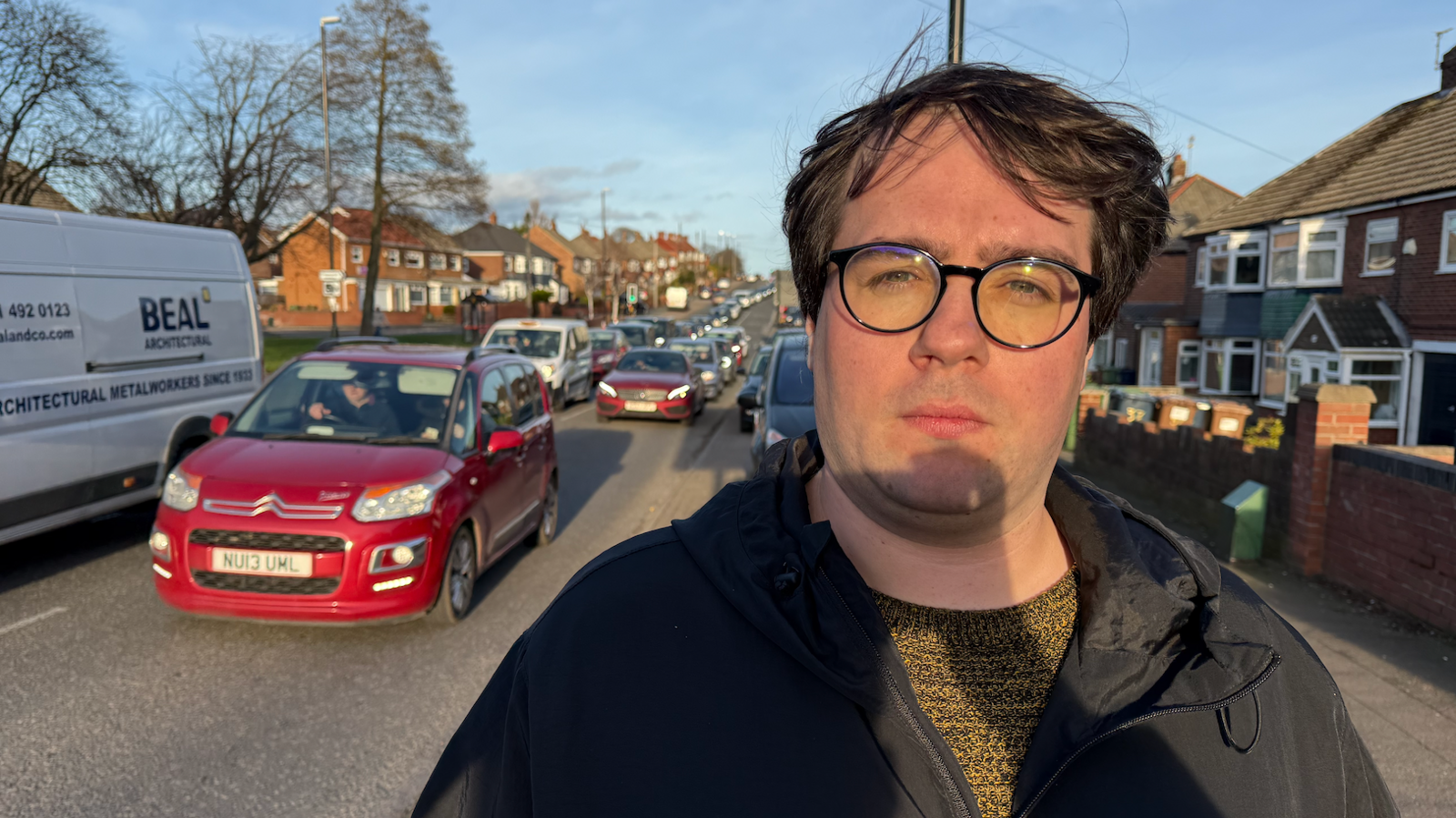 A man with brown hair and wearing thick black rimmed glasses stands next to a busy road where there is a queue of cars. 