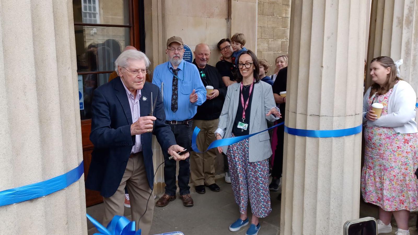 A white-haired man in his 80s, wearing a dark blue jacket, a light blue shirt and light blue trousers, holds up a pair of scissors having just cut through a ceremonial blue ribbon tied between two columns at the ornate entranceway into a public building. Various other people stand around watching and applauding.