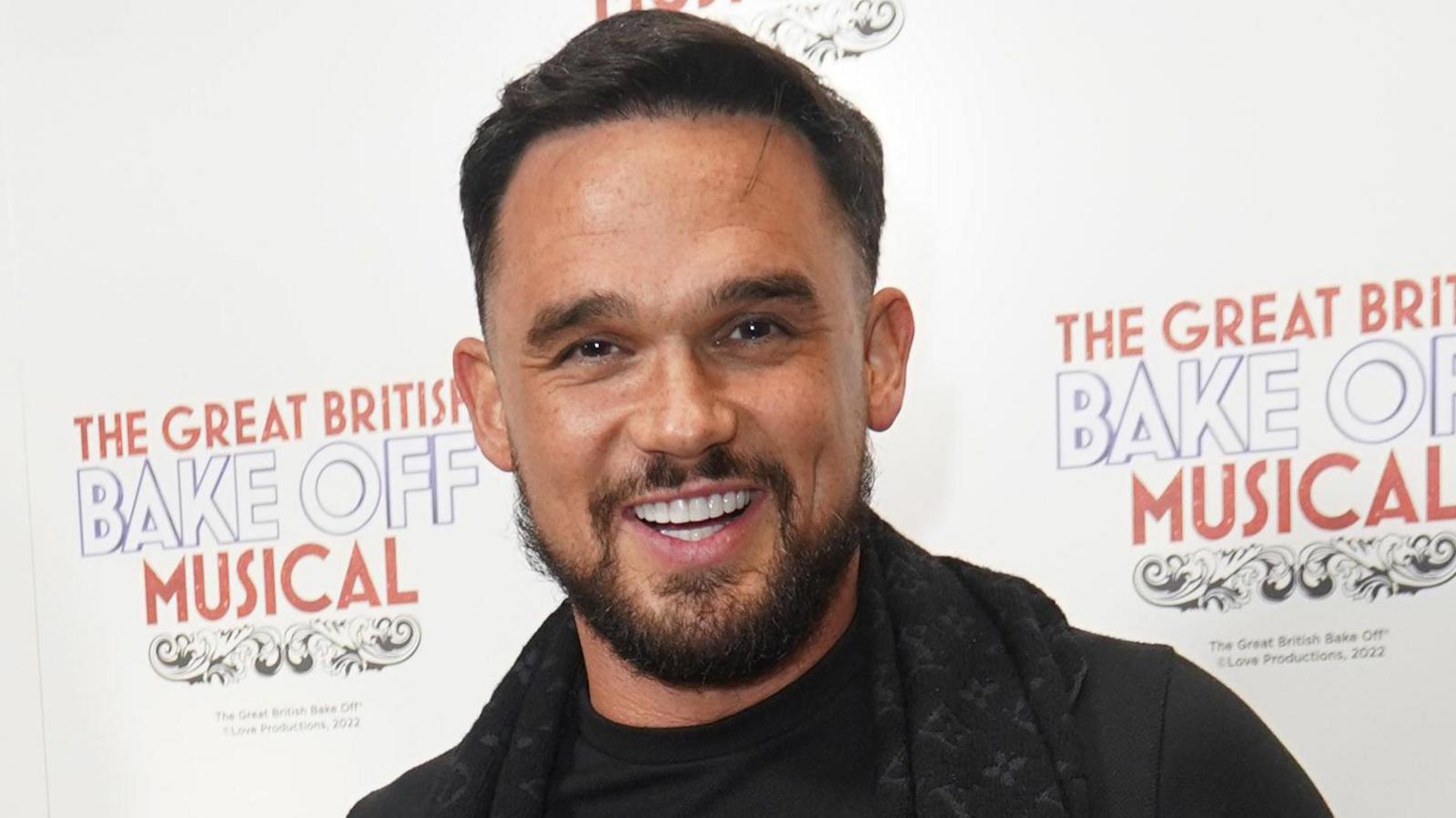 Gareth Gates smiles at the camera. He has short brown hair and a beard and is wearing a black T shirt and black Louis Vuitton scarf. 