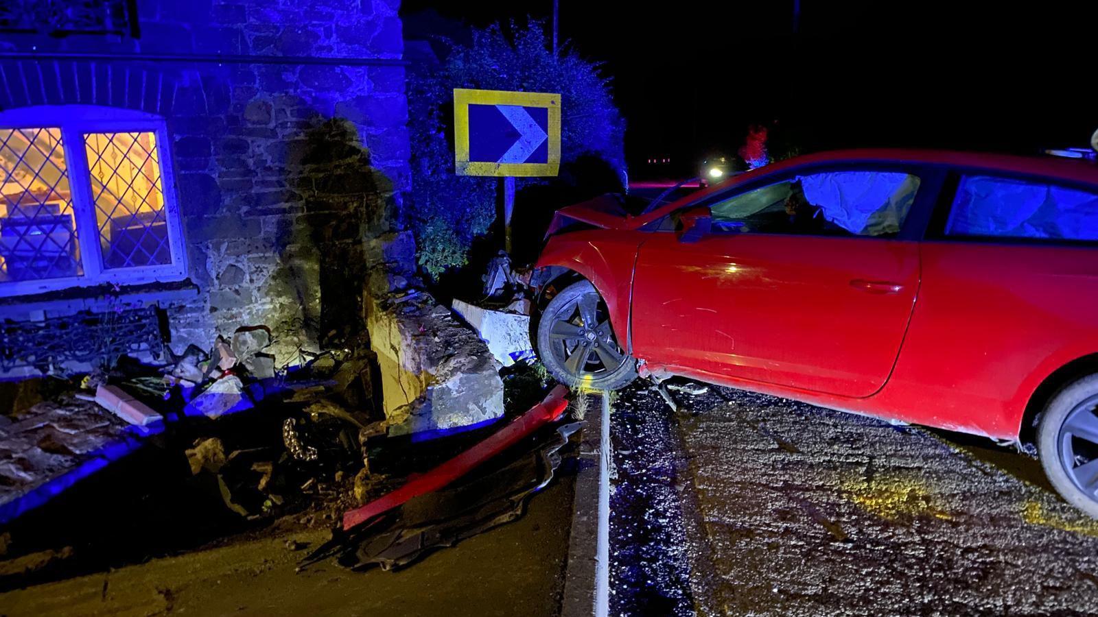 In the dark, a red car has crashed on the side of the road. Bricks from a cottage based house have been knocked over by the car. Yellowish light is coming from a glass window. There's been pieces from the front of the car that have detached and left on the ground. An arrow on a sign that's facing towards the right is opposite to the car. 
