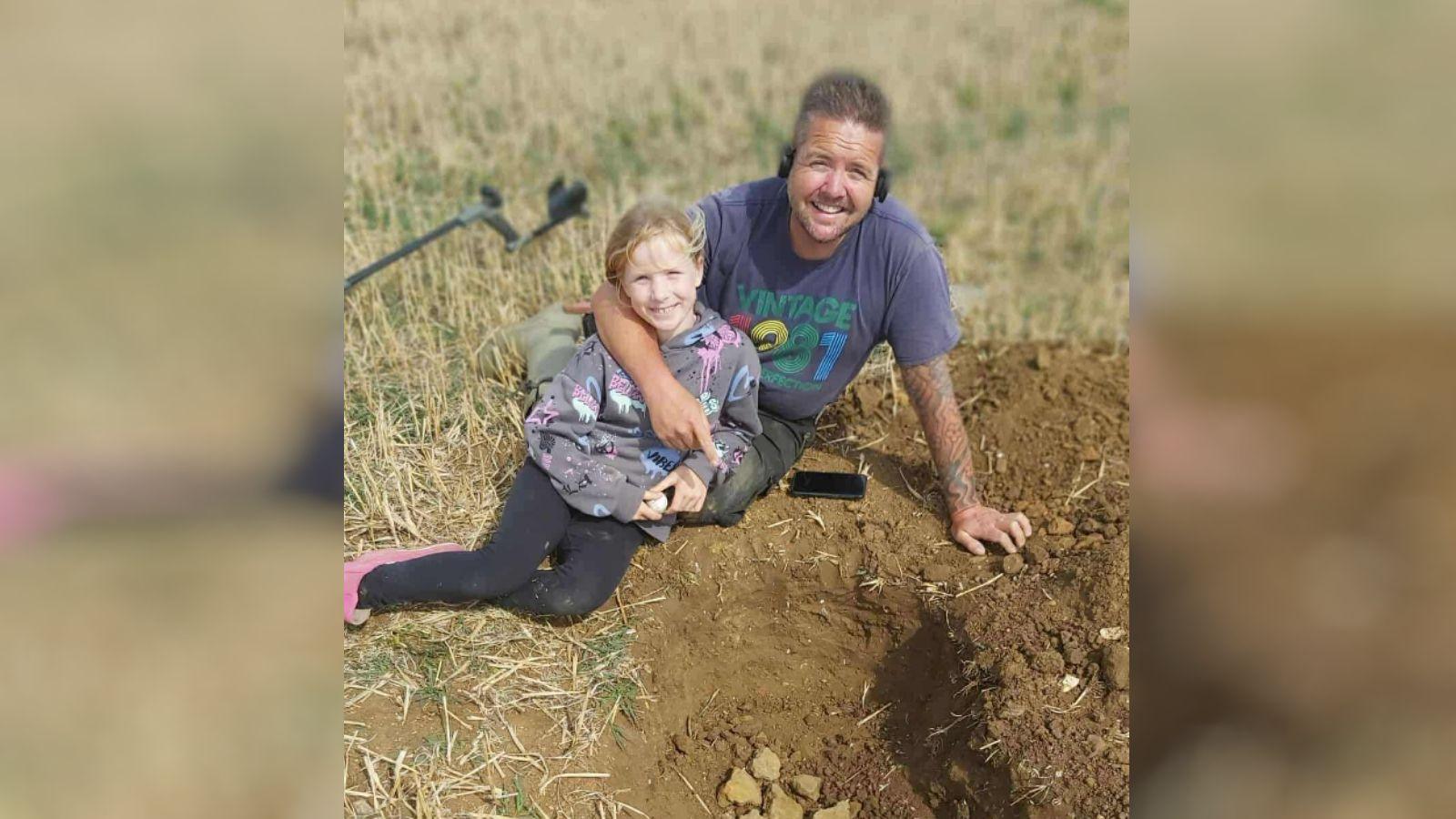 A man and his daughter lie on a field. In front of them is a dug hole that has some lumps of metal inside. 