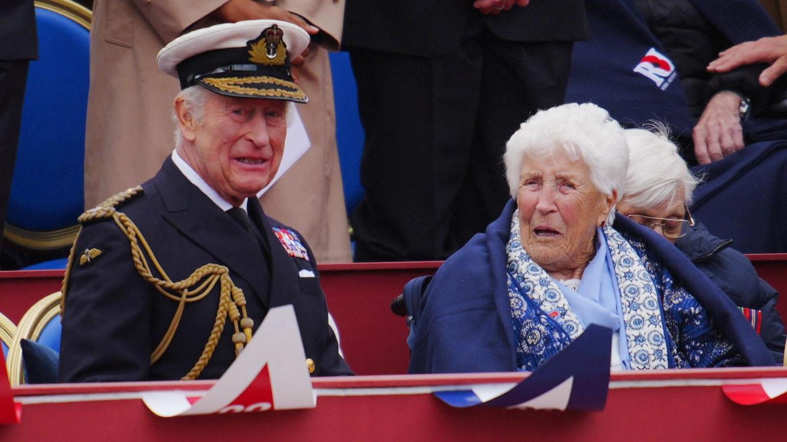 King Charles sits in full military uniform in the box during the VE day military parade - Joy Trew sits next to him wearing a blue patterned scarf and wrapped in a navy blue blanket