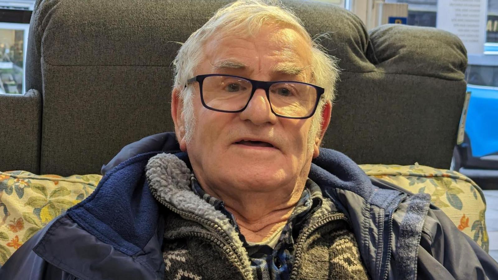 A man wearing glasses, a navy blue coat and a pullover sitting on a yellow and blue patterned sofa.