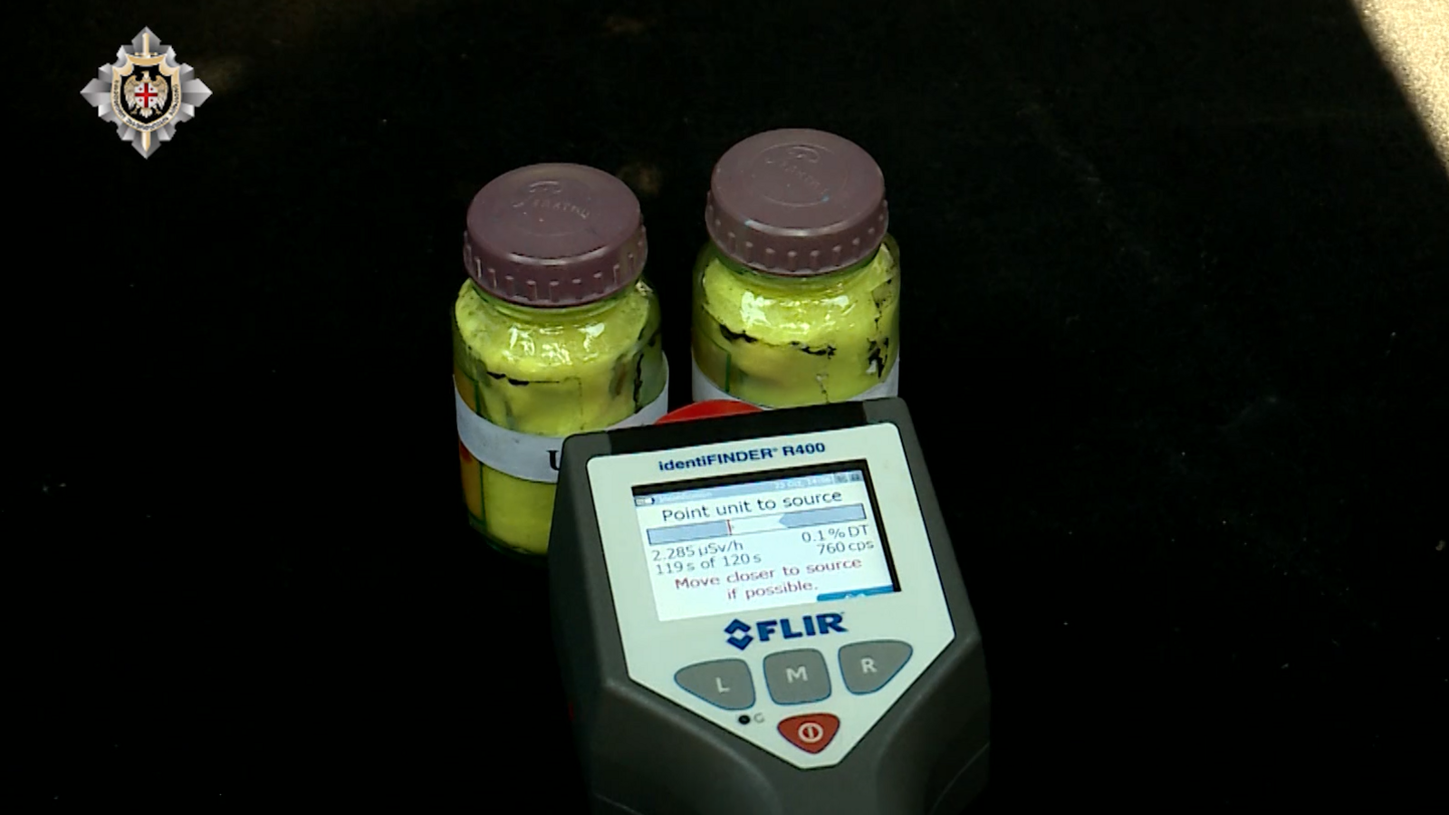 Two glass jars containing a yellow substance in a car boot with a dosimeter pointing at them.