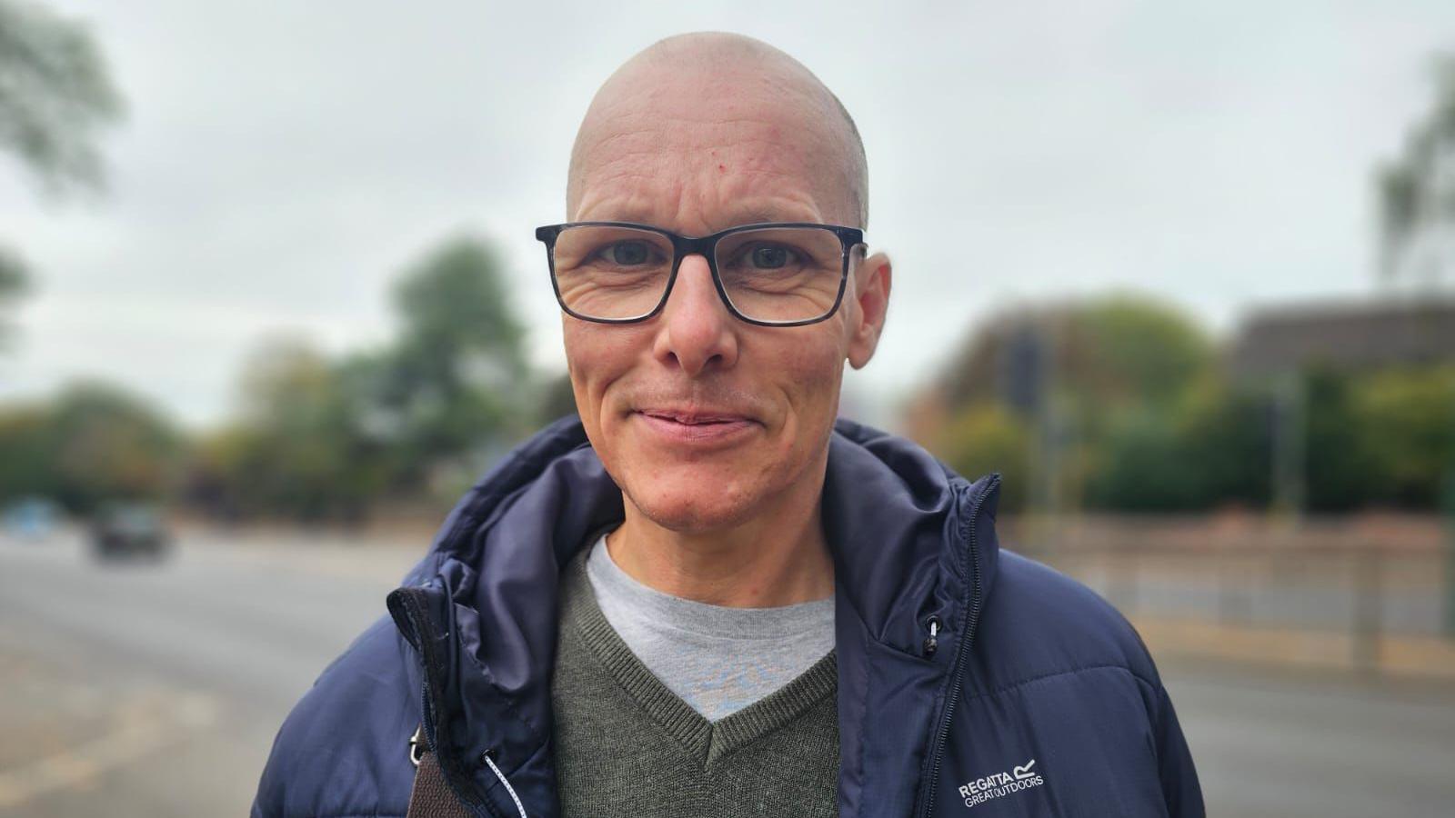 Mr Callan is smiling at the camera, he has black framed square glasses and a bald head. He is wearing a grey t-shirt and sweatshirt and a navy hooded coat.
