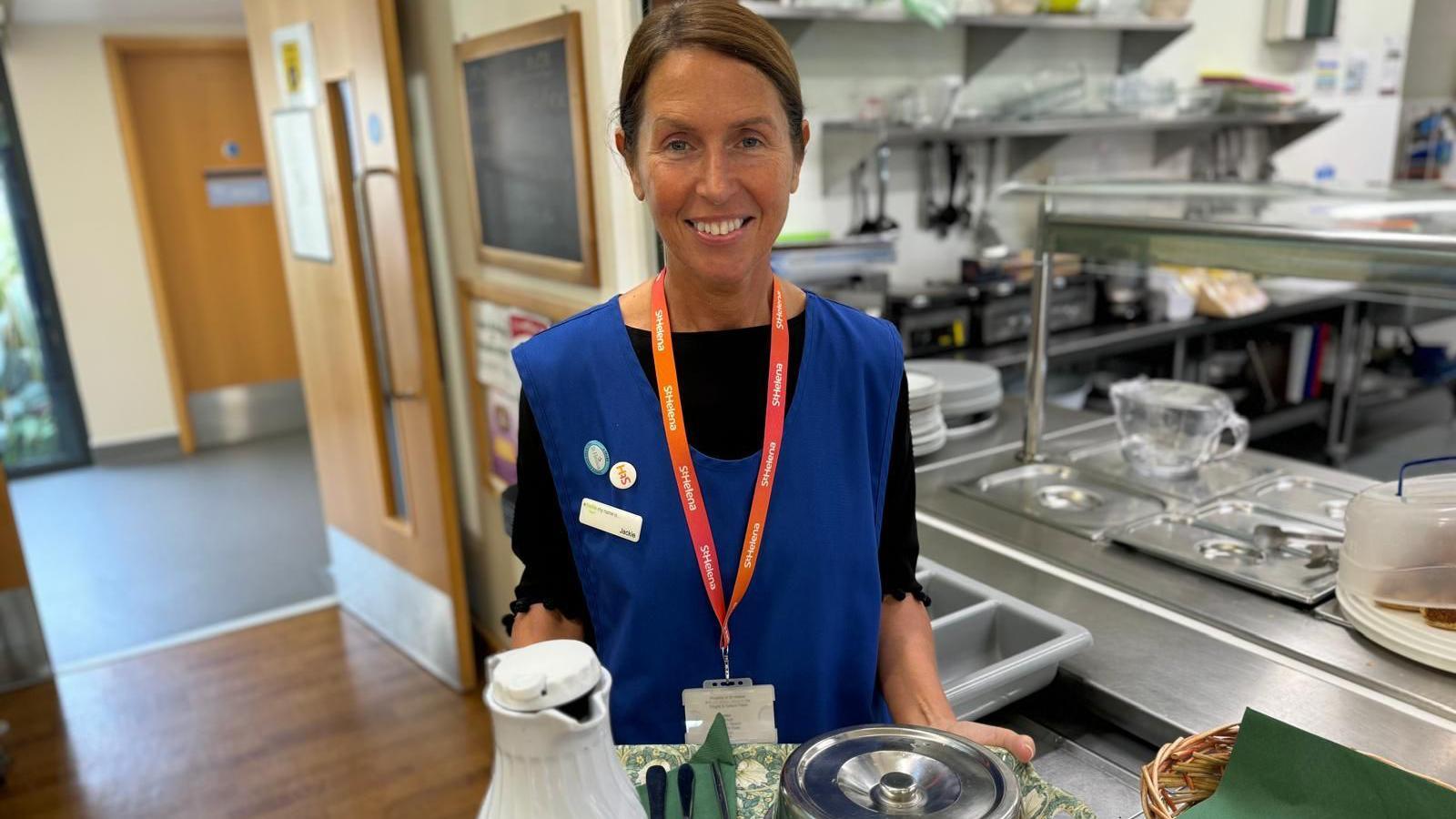 A smiling woman holding a tray, with a jug at a metal plate cover on it, in front of a commercial kitchen. She is wearing a blue tabard and an orange lanyard.