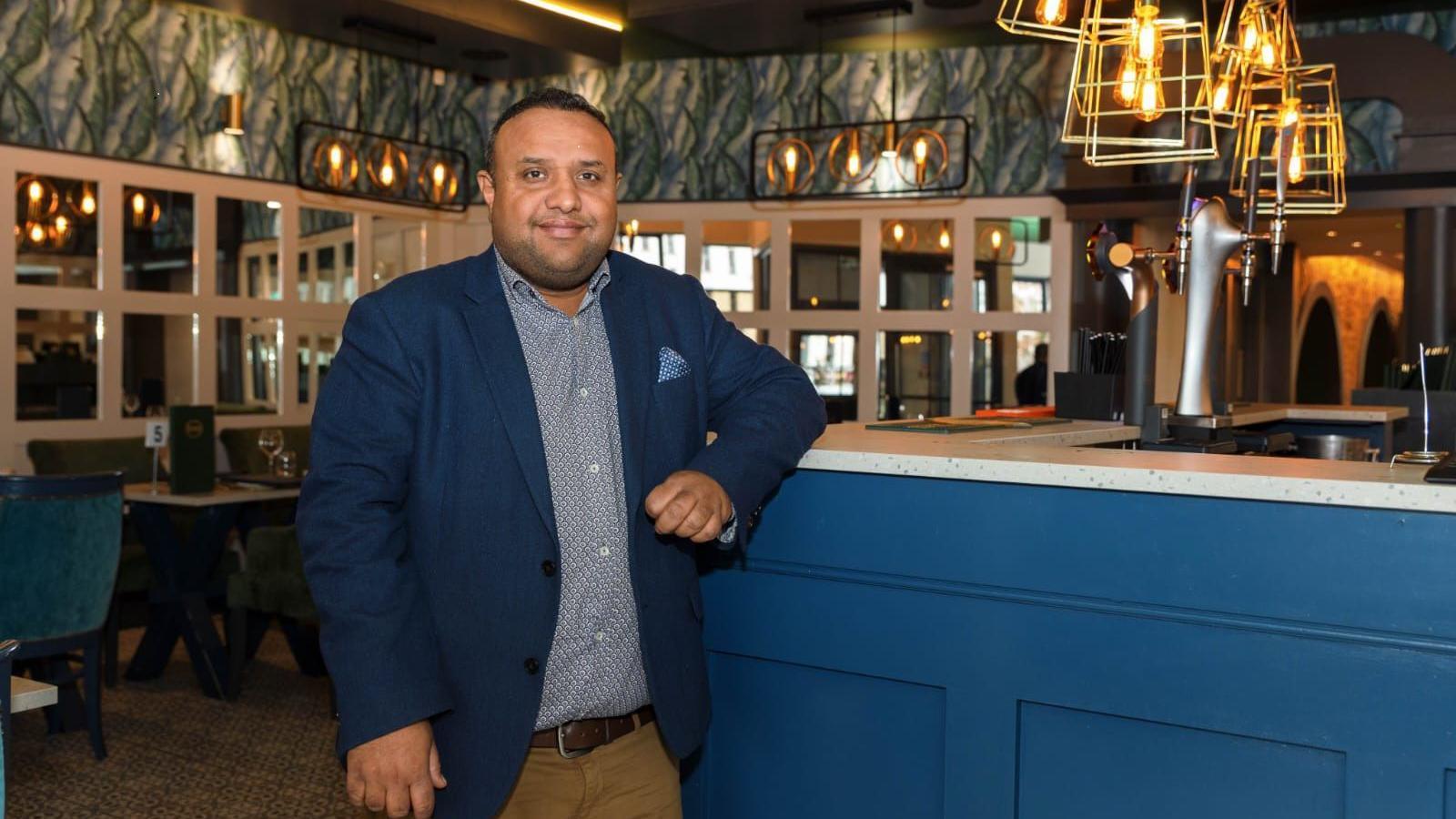 Gaurav Barot in his restaurant in Newry.  He has short, dark hair and is wearing a navy blazer, a blue patterned shirt and beige trousers with a brown belt.  