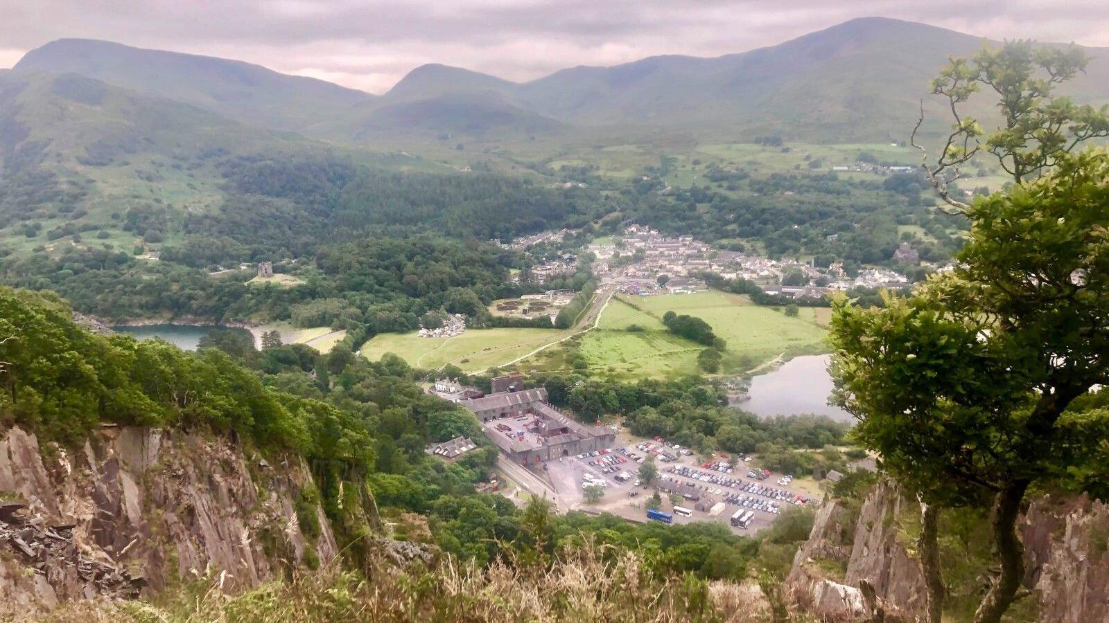 View of the National Slate Museum from above, taken from the top of the nearby Vivian Quarry, looking down on the museum and the green of the valley it sits in, with a backdrop of mountains beyond