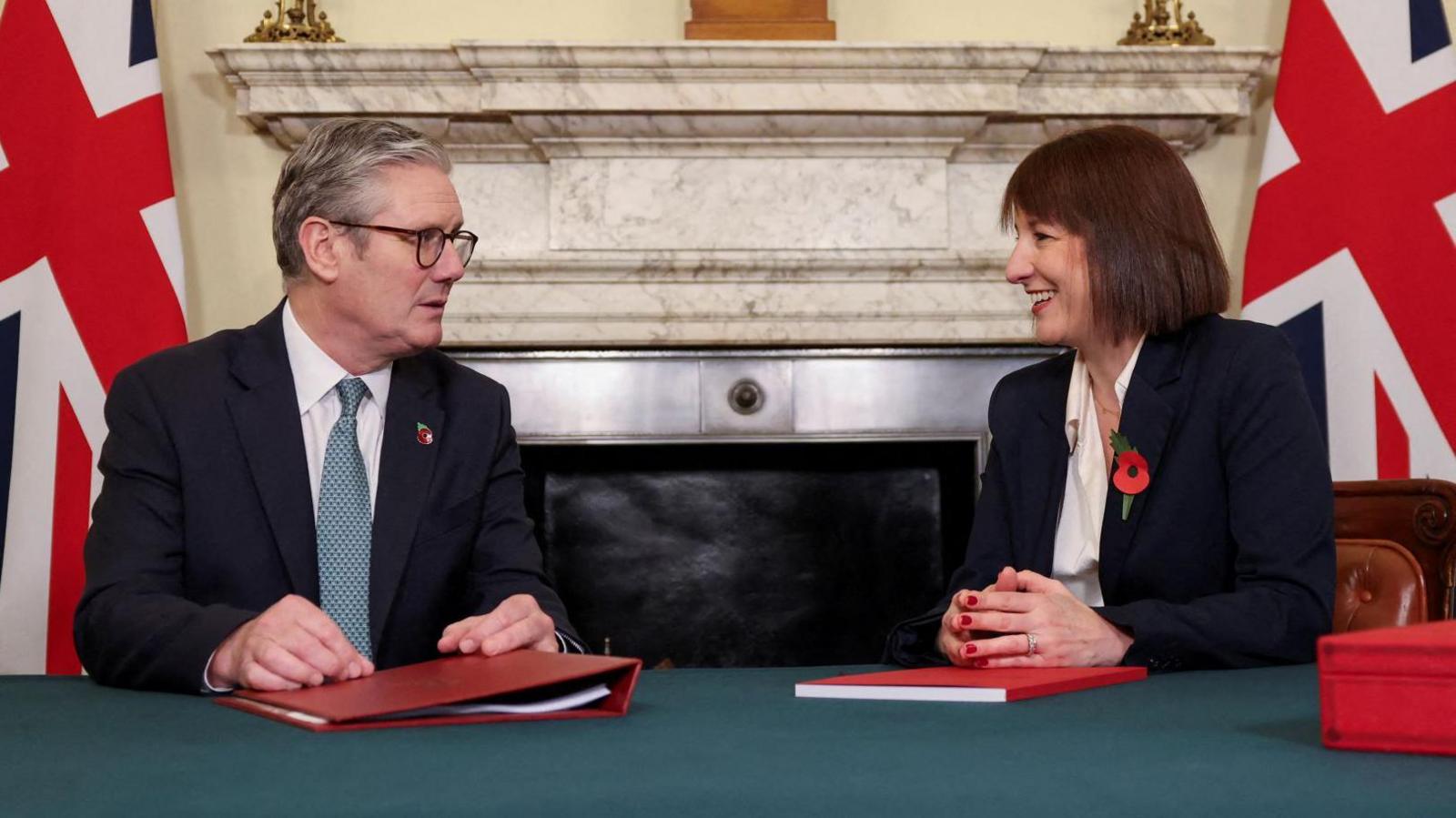 Sir Keir Starmer and Rachel Reeves sit at a table with red binders and notebooks in front of them. They are in front of a large fireplace with British flags either side of it. 