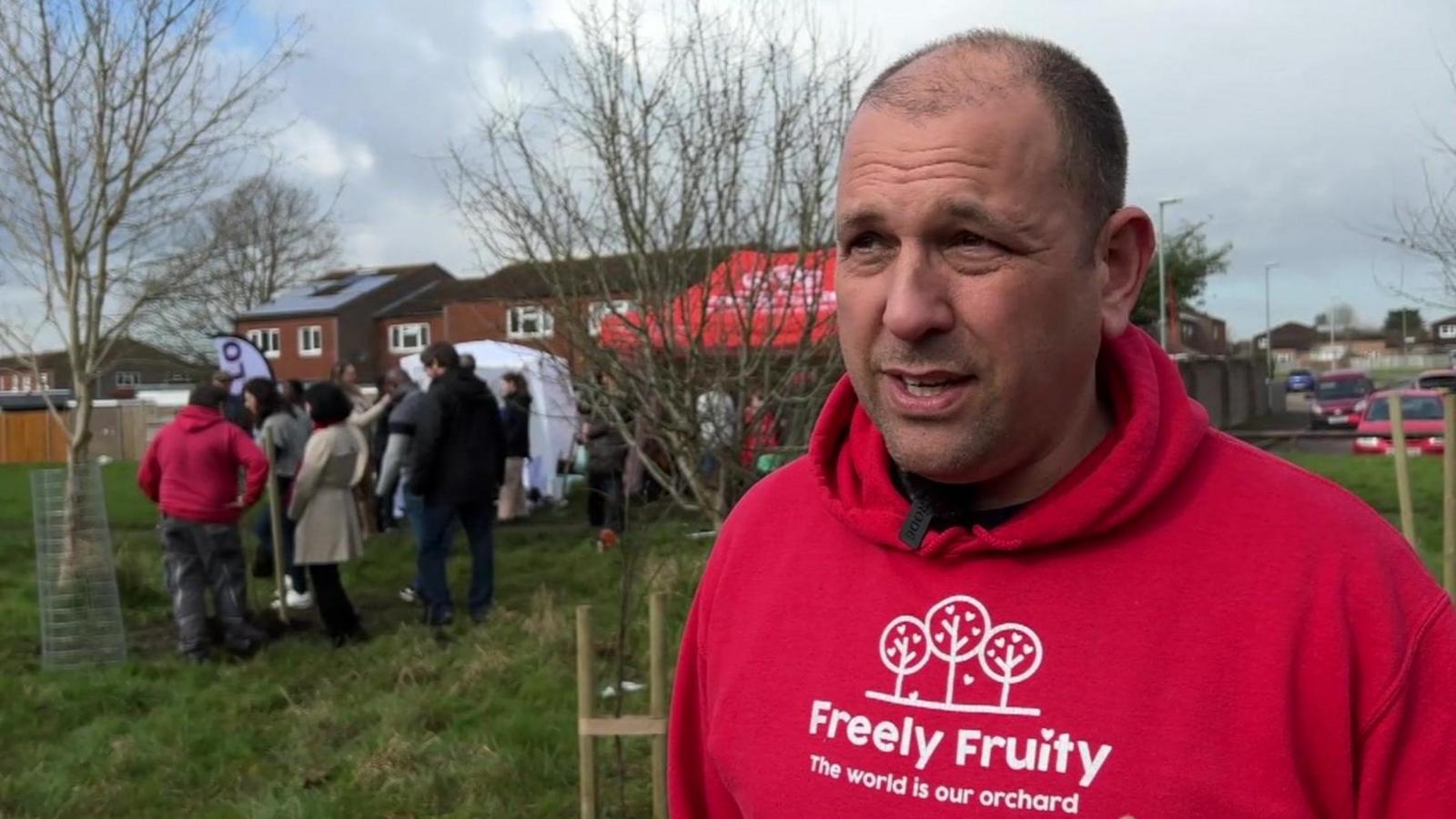 Matt wears a red Freely Fruity hoodie with the motto 'The world is our orchard'. Tree planting is happening in the background.