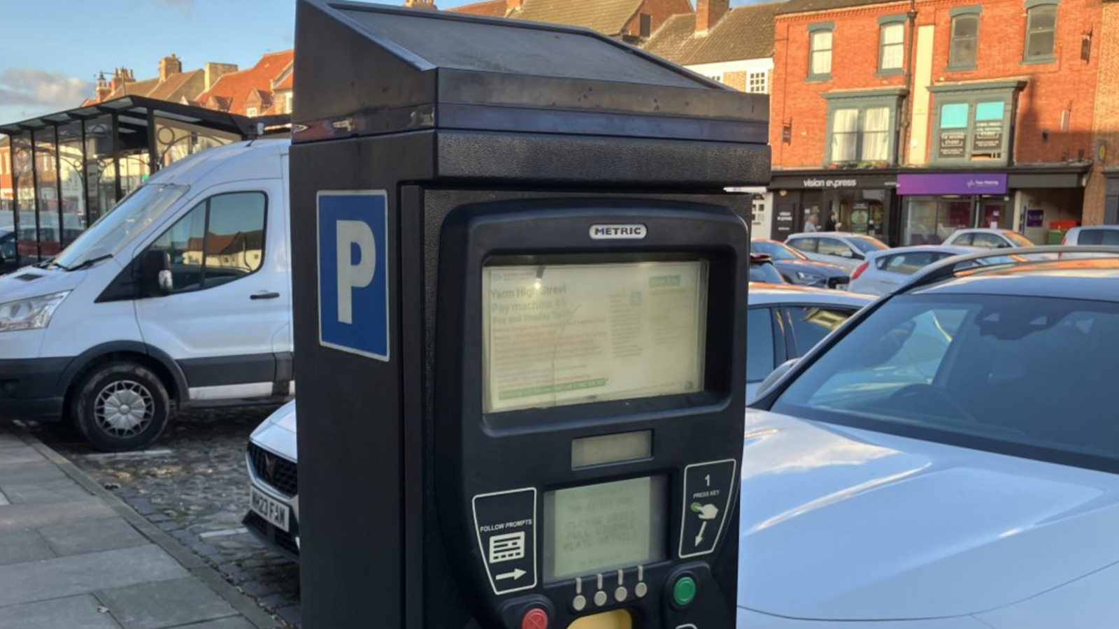 A parking meter with two cars and a van behind it and shops in the background.