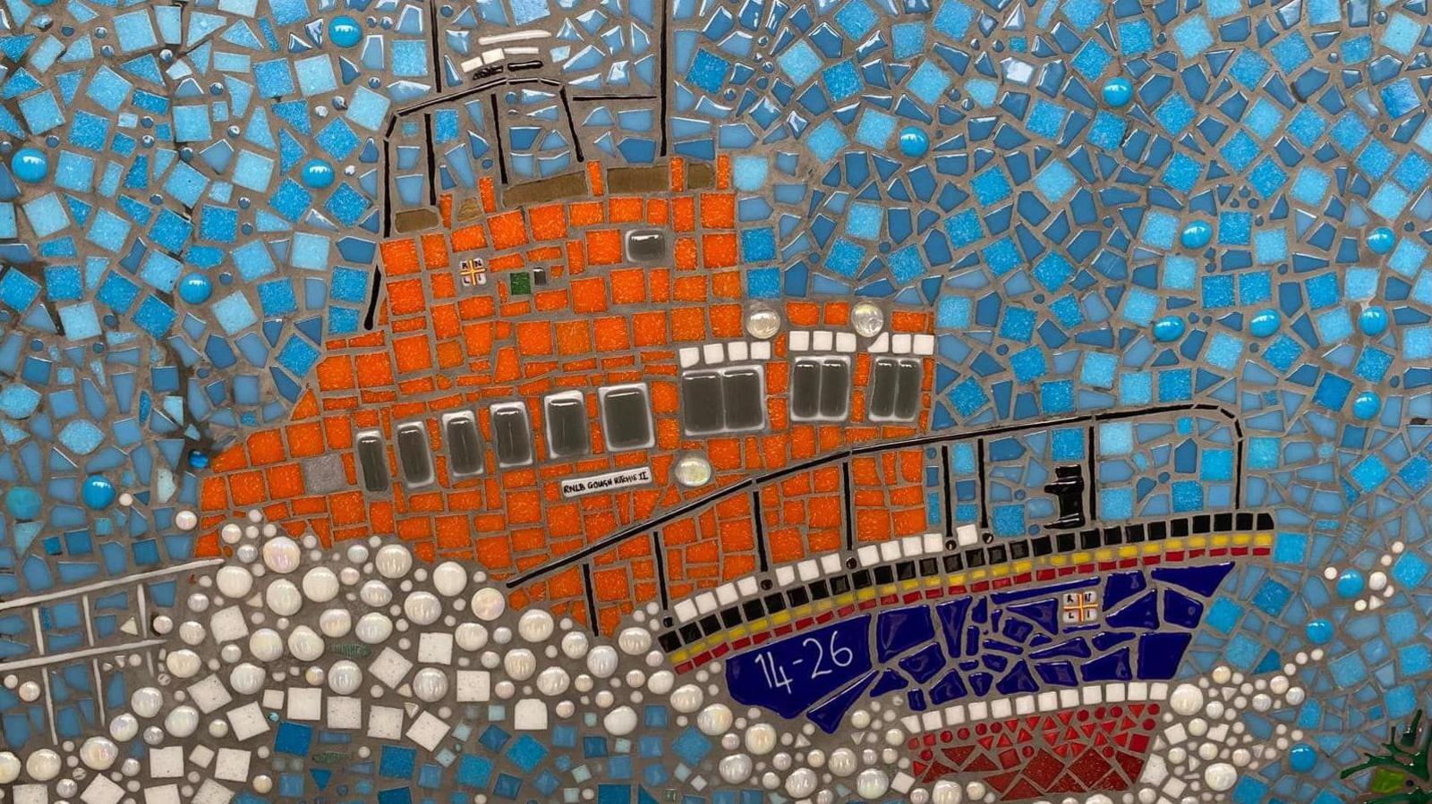 Part of the mosaic featuring the Port St Mary lifeboat in the RNLI colours of orange with a blue and red hull that also features bands of black, yellow and white. It's set against blue and white tiles representing breaking waves.