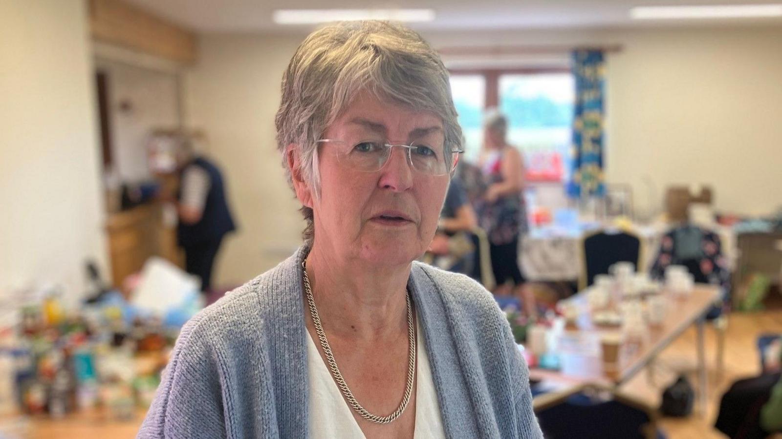 A grey-haired woman wearing frameless glasses and a grey cardigan, standing in front of a blurred jumble sale, inside a cream-walled community centre