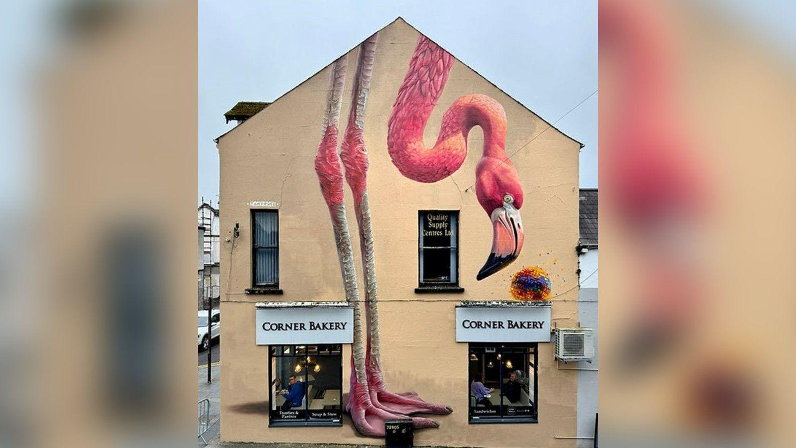 An example of a mural by Blank Wall Assassins' artist Woskerski. It is a flamingo bent down to look at a colourful object. It is painted on the side of a building with the sign "Corner Bakery" above its two windows.