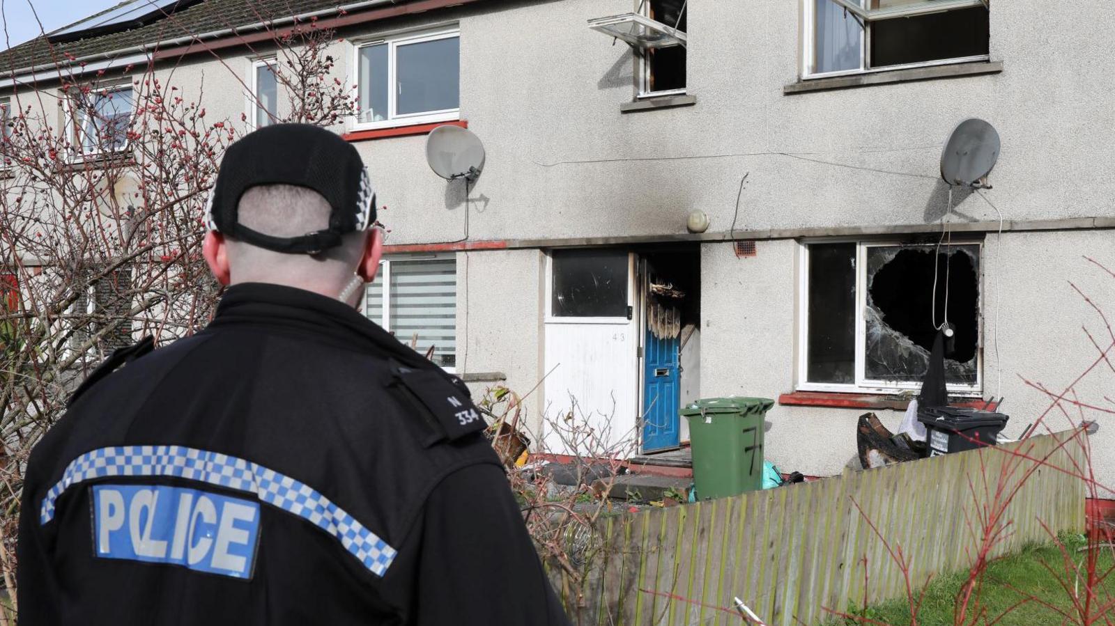 A police officer, with his back to the camera, is looking at the house. The top windows are open wide, while a door and a window on the ground floor show signs of fire damage. The ground floor window is also broken.