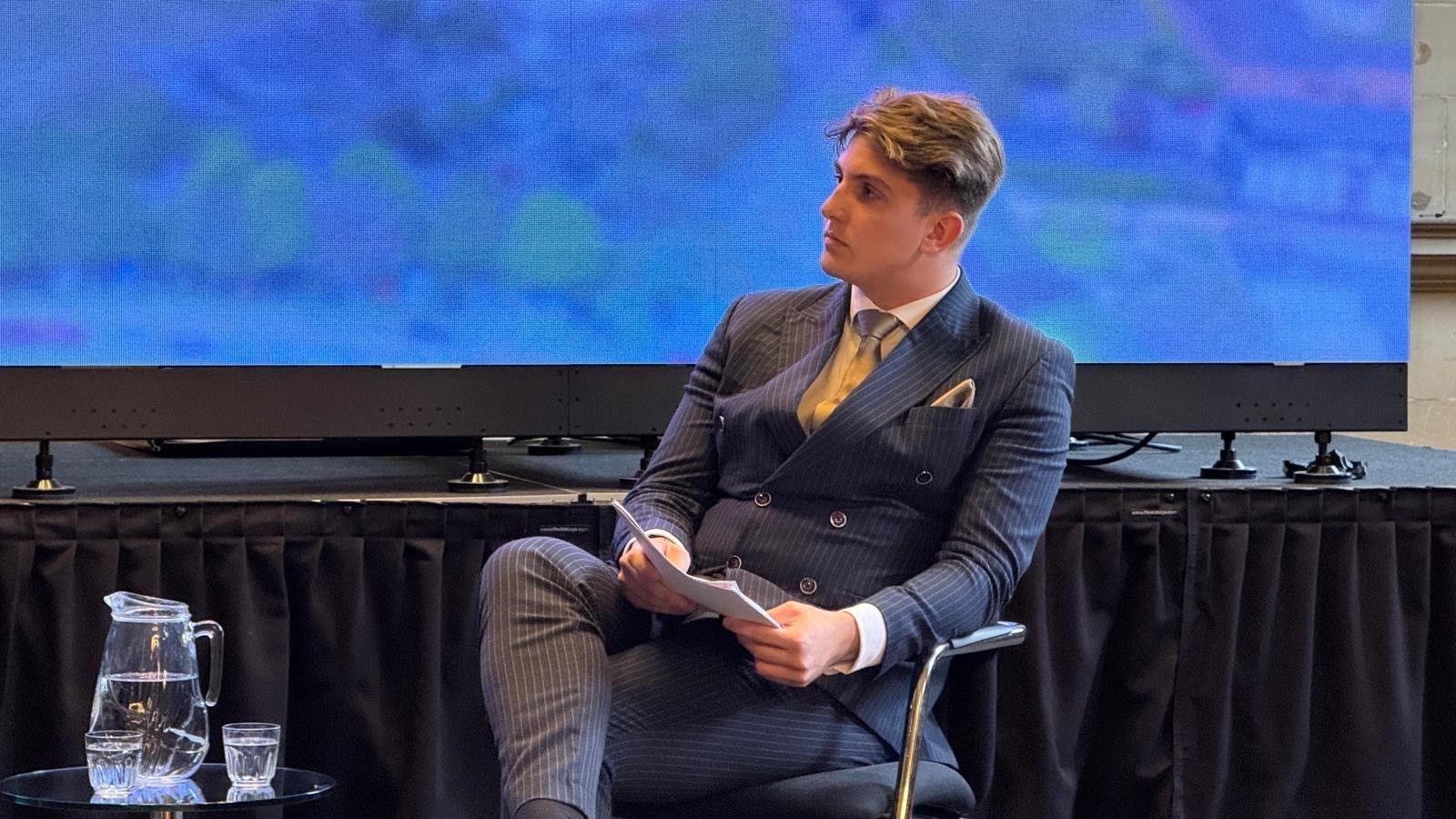 A man sitting down in front of a low stage in a navy pinstripe suit.