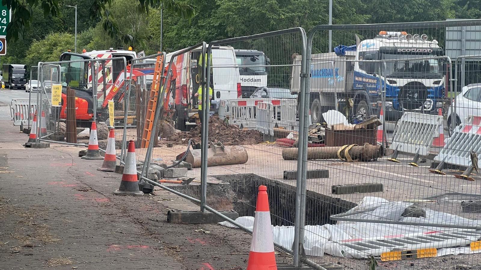 A fenced off hole in the ground with cones around it and traffic passing by behind it