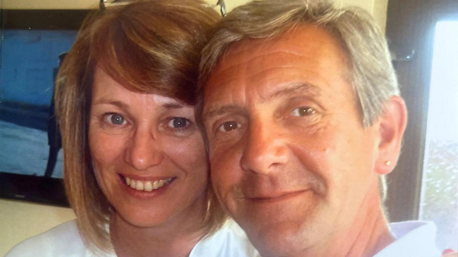 Christine and Stuart Cullen are smiling looking at the camera. They are both wearing white tops. Christine has her glasses on her head.